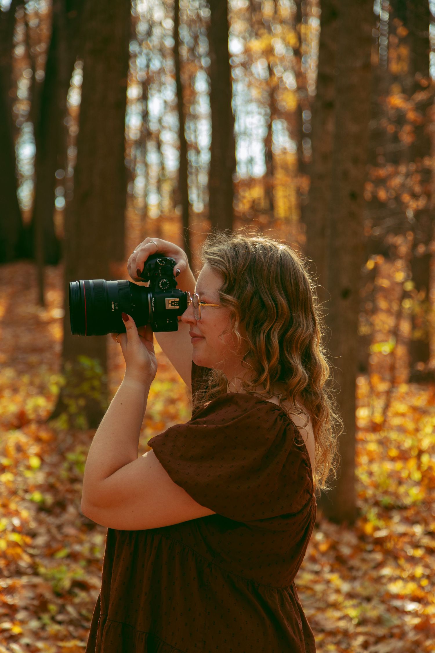 Woman in a brown dress and glasses holds a camera to her face, photographing in a sunlit autumn forest with golden fallen leaves.