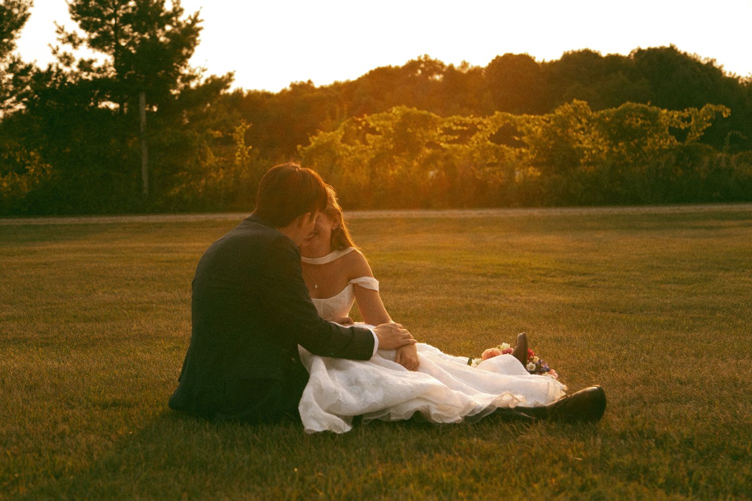 Bride in a white gown and groom in a dark suit sit close together on a grassy lawn at golden hour sunset, framed by trees.