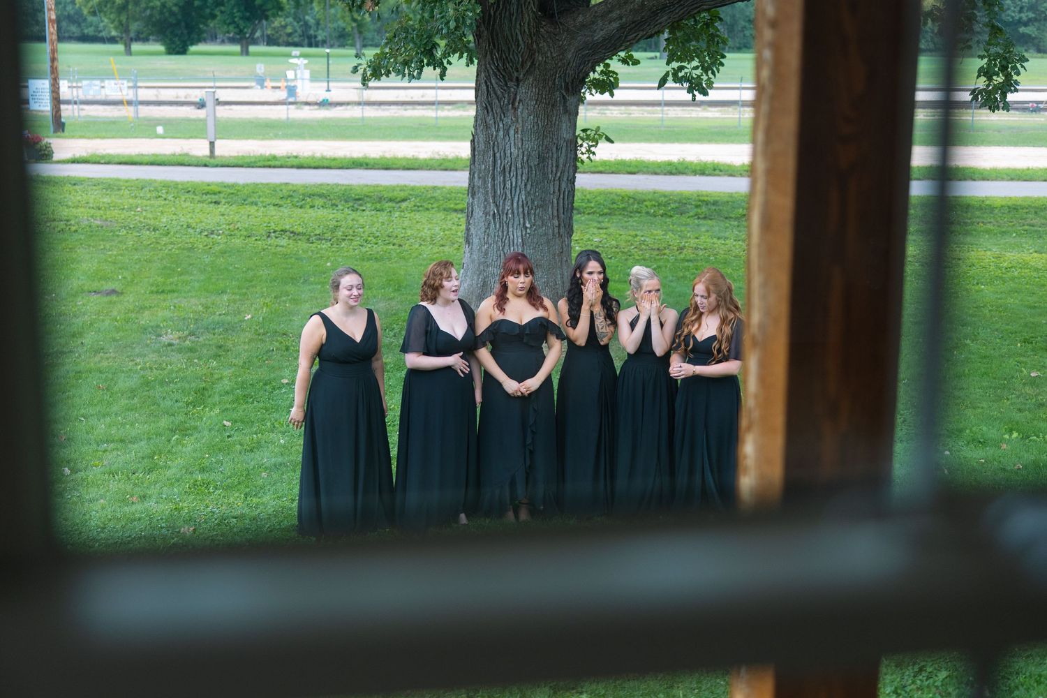Six bridesmaids in long black dresses stand together on a green lawn beneath a large tree, viewed through a window frame.