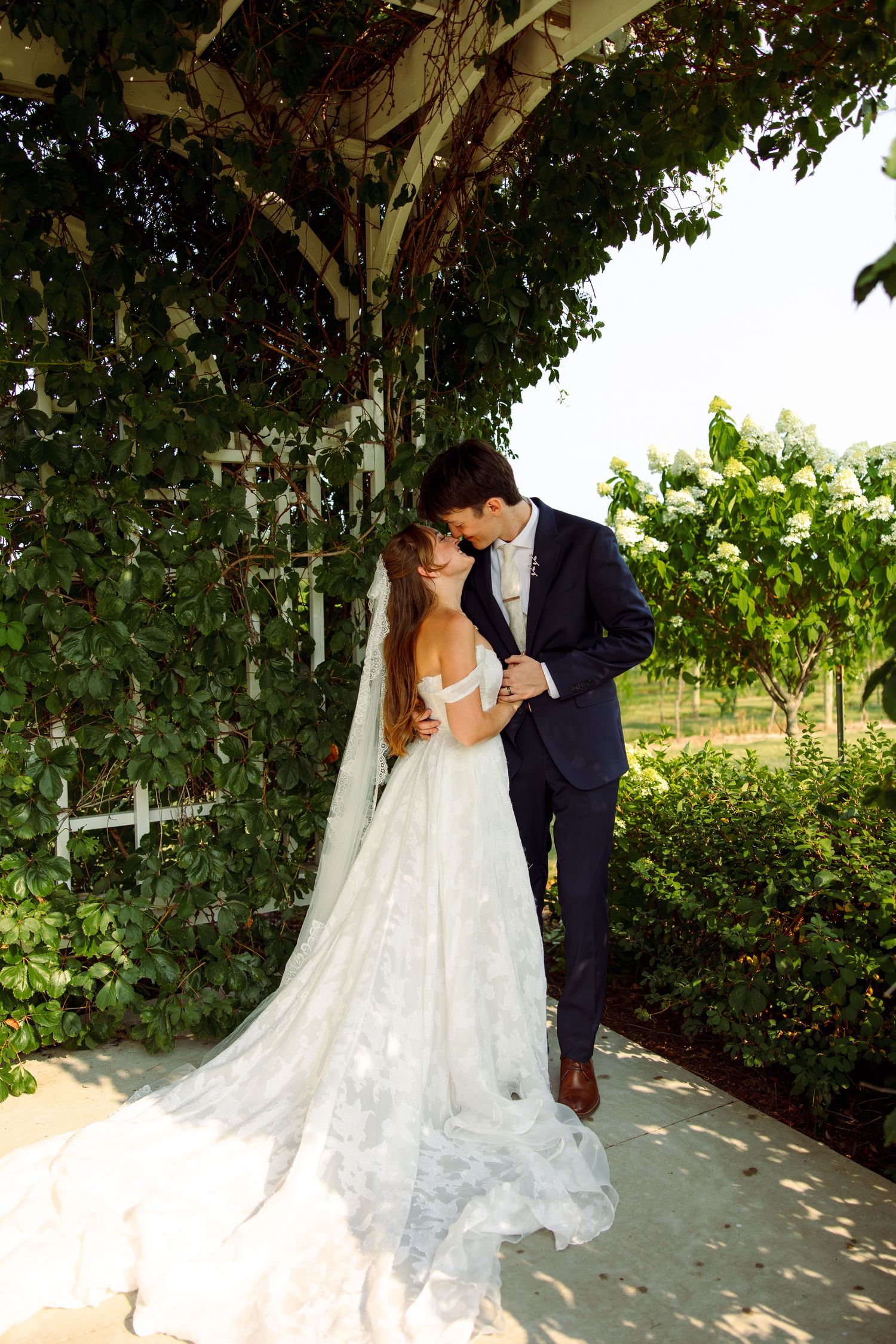 A groom in a navy suit leans in to kiss his bride in a lace wedding gown with a long train beside a leafy trellis.