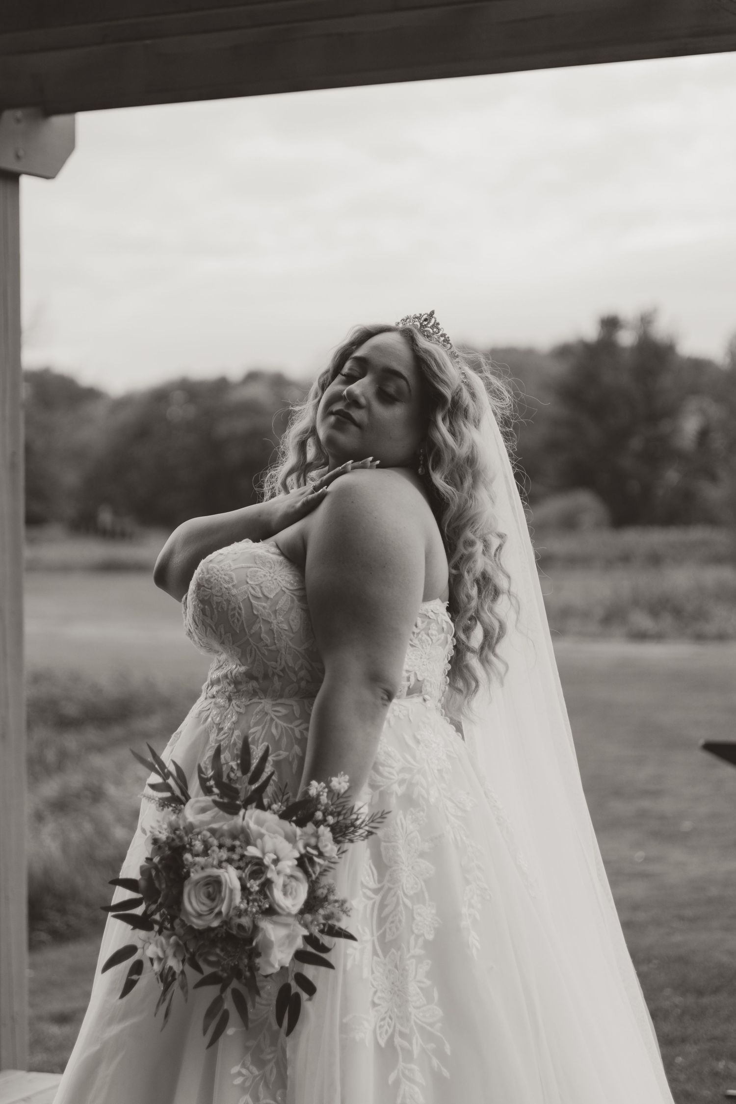 Black and white photo of a bride in a lace wedding gown, veil and tiara holding a floral bouquet and posing outdoors by a field.