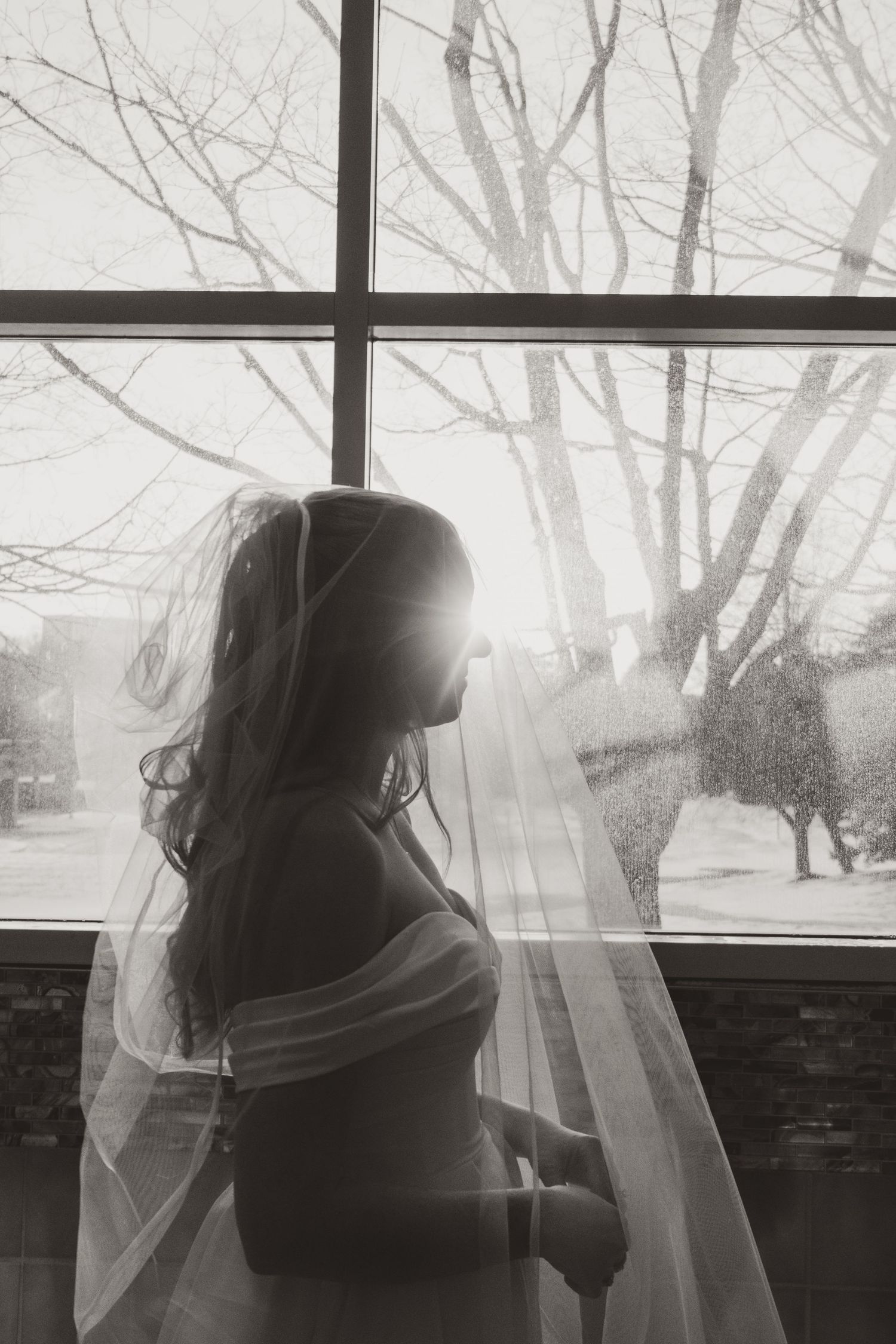 Black and white portrait of a bride in a sheer veil and off-the-shoulder gown gazing out a sunlit window.