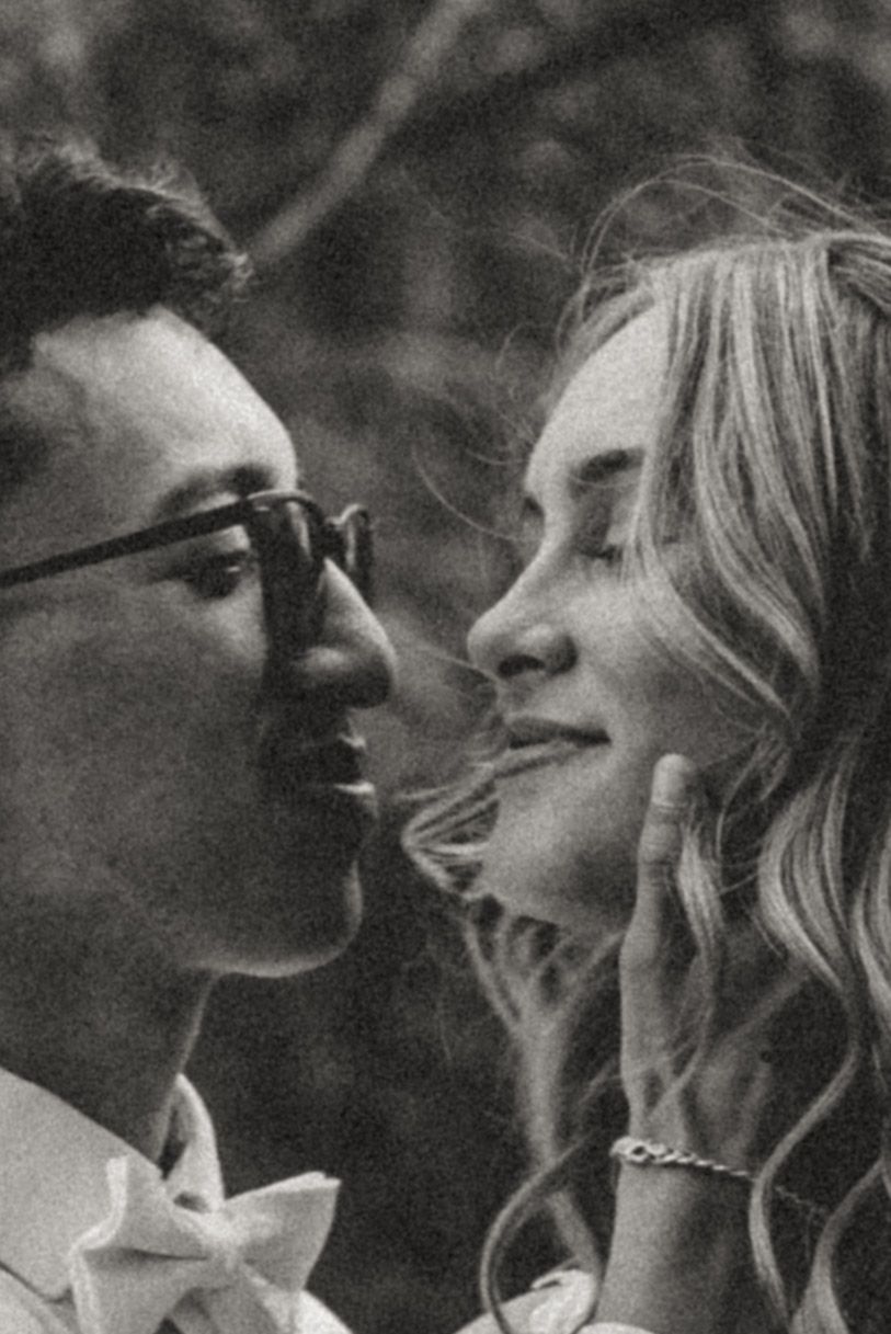 A black and white close-up of a groom in glasses and a bride with wavy hair about to kiss as she touches his face.