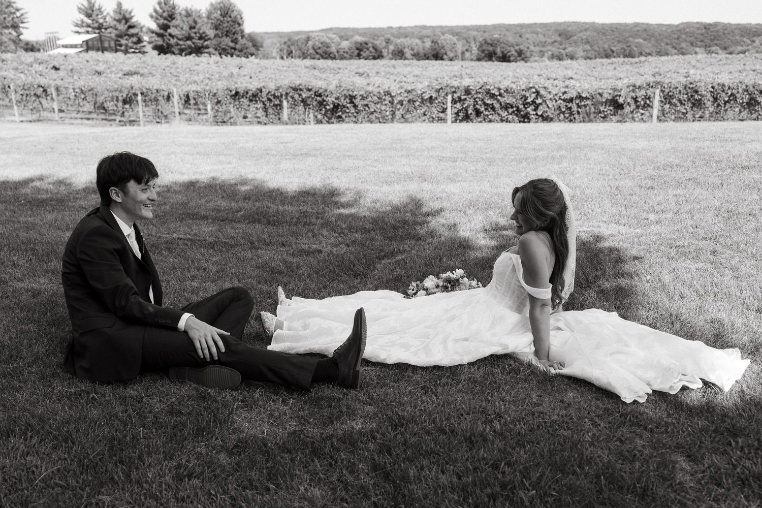 In a black and white photo, a bride in a flowing gown and a groom sit facing each other on the grass in front of a vineyard.