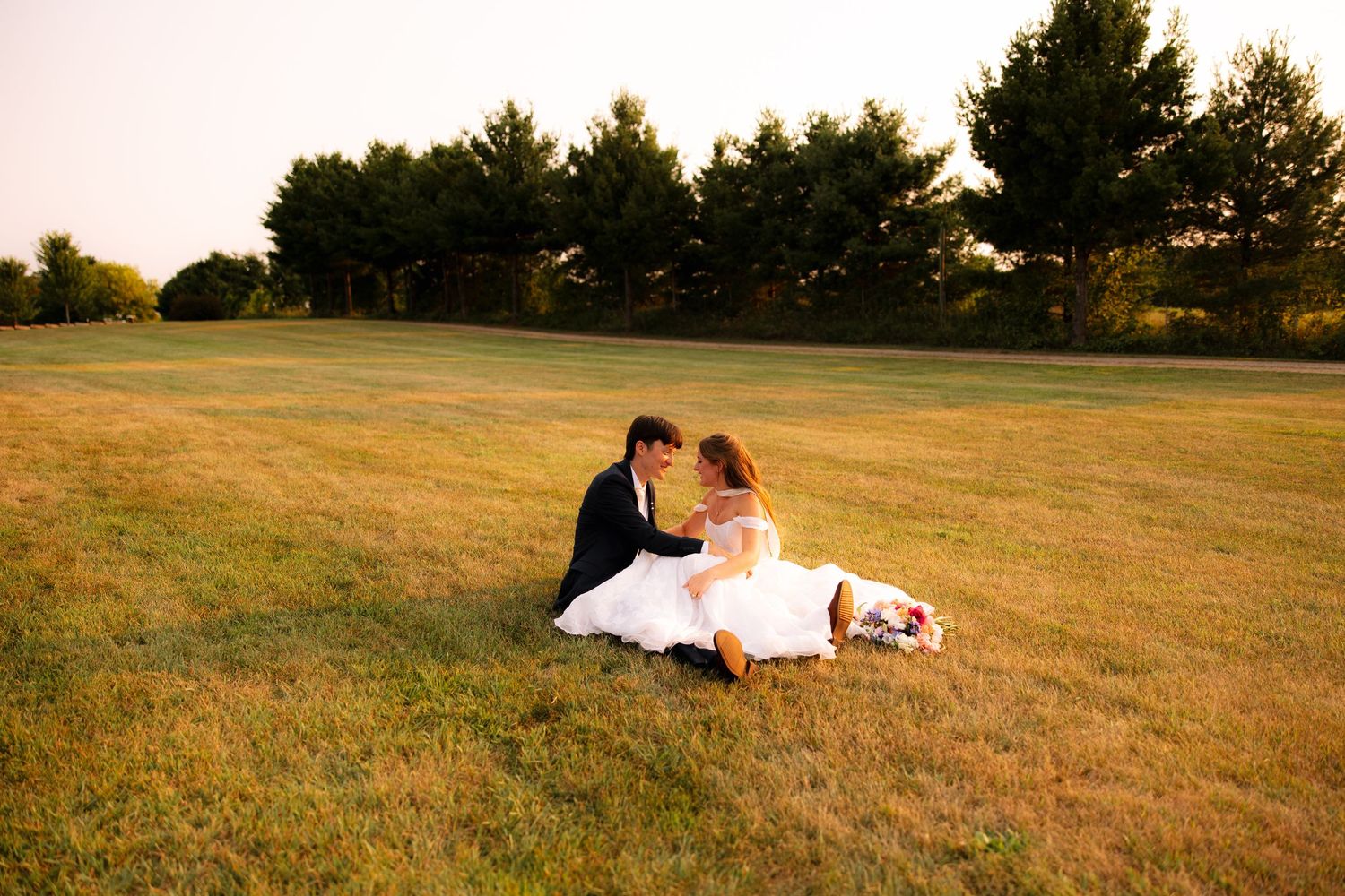Bride in a white dress and groom in a dark suit sit close together on a wide grassy field at sunset, surrounded by tall trees.