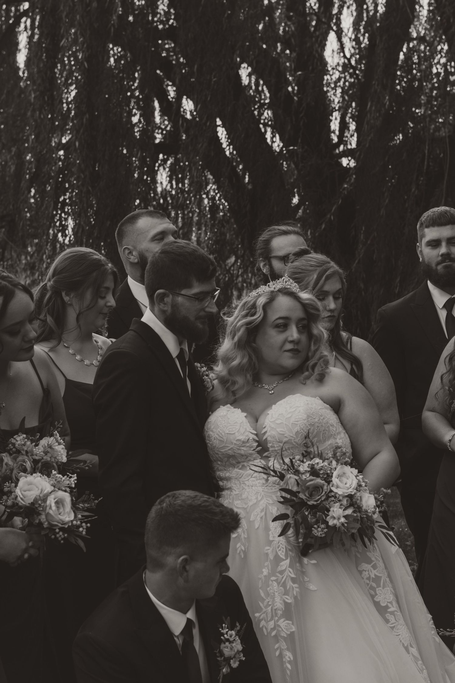 Black and white portrait of a bride and groom surrounded by their bridal party holding bouquets beneath weeping willow branches.