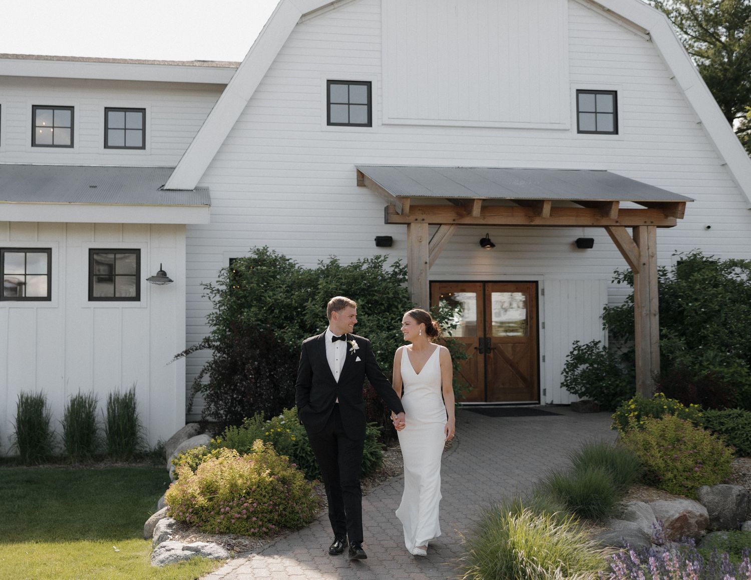 Bride and groom walk hand-in-hand outside a white barn wedding venue surrounded by lush greenery.