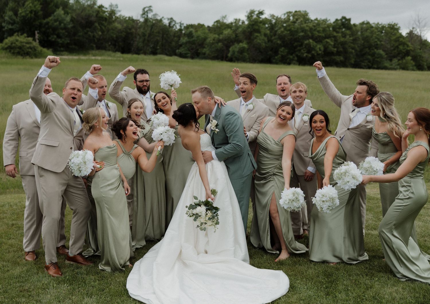 Bride and groom kissing surrounded by cheering wedding party in sage green and tan attire on a lush green lawn.