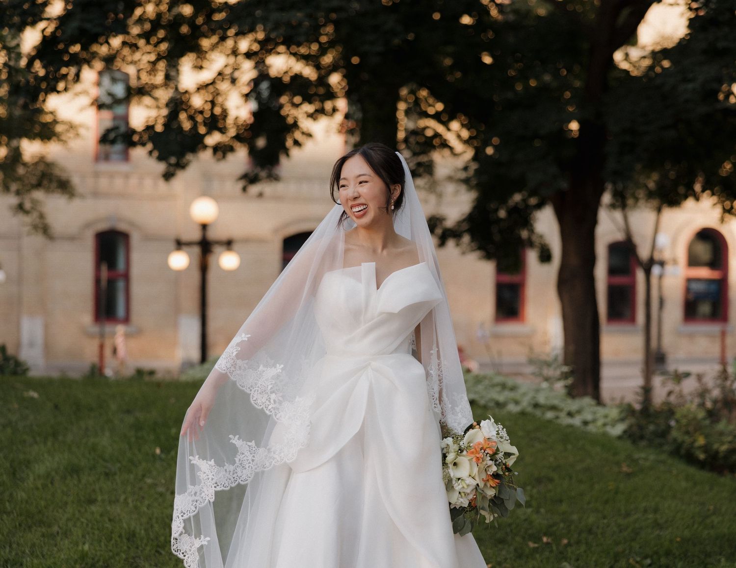 A bride in a flowing white wedding gown holds her veil while walking through a garden at sunset.