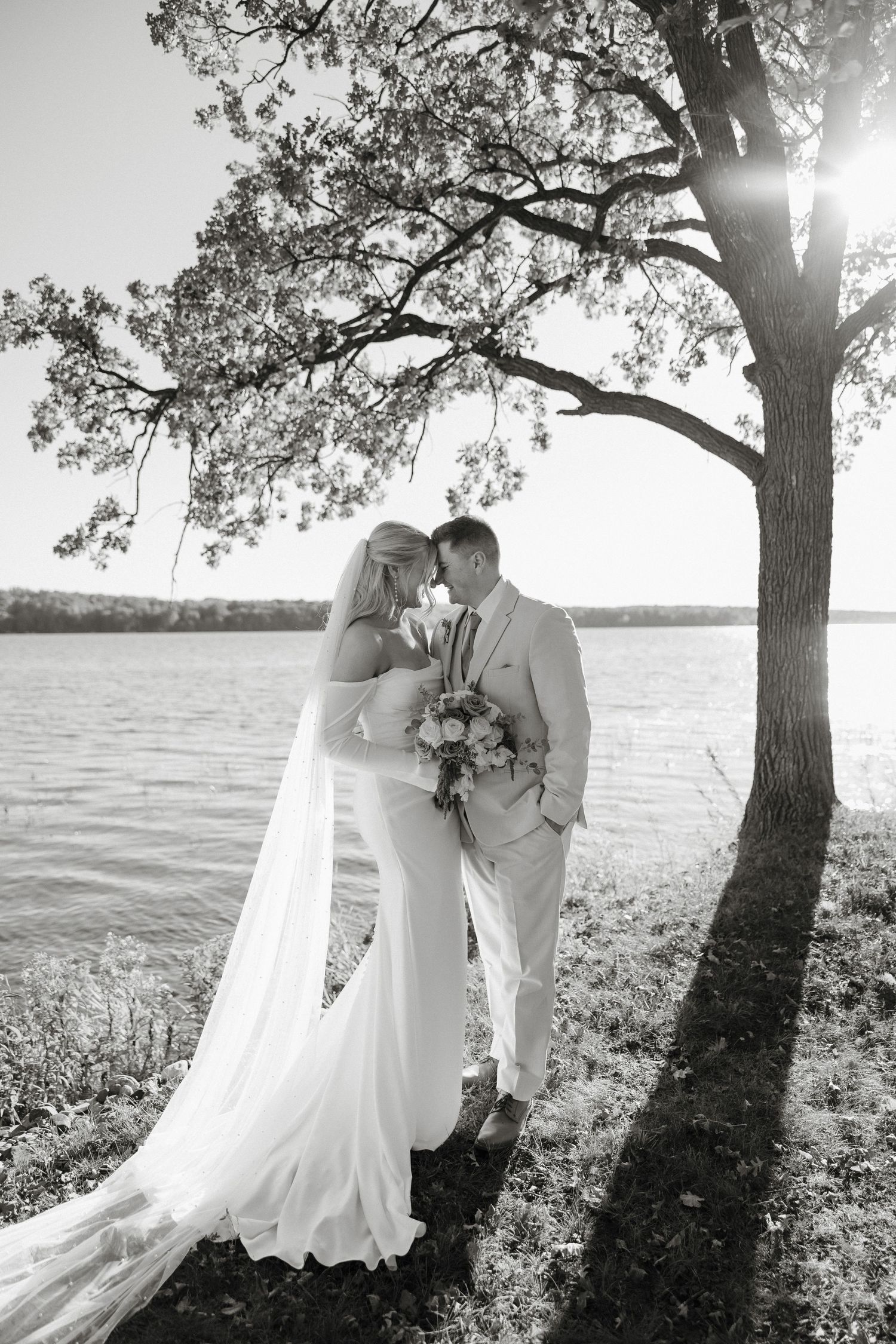 Romantic black and white wedding photos of a couple by a lake under a tree with sunlight filtering through branches.