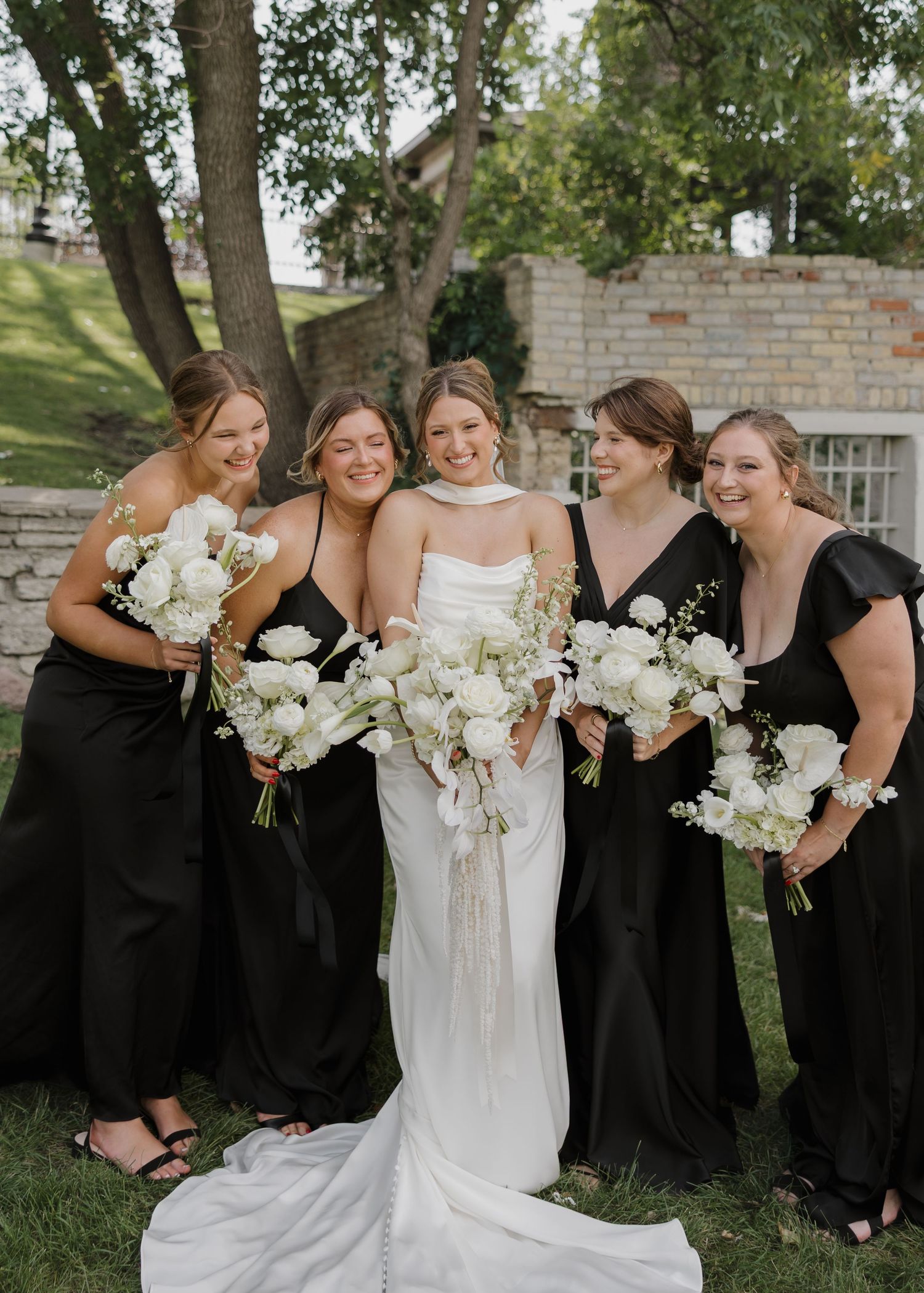 Wedding party in elegant black bridesmaid dresses and white gowns pose together outdoors with white floral bouquets.