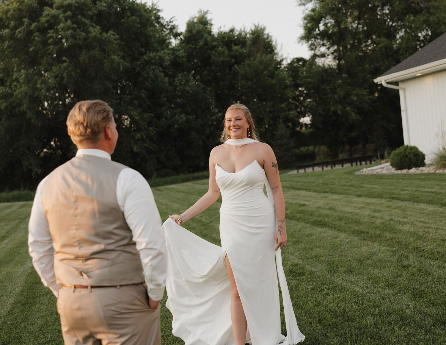 A bride in a white fitted wedding dress walks on a manicured lawn with someone in a tan vest on a summer evening.