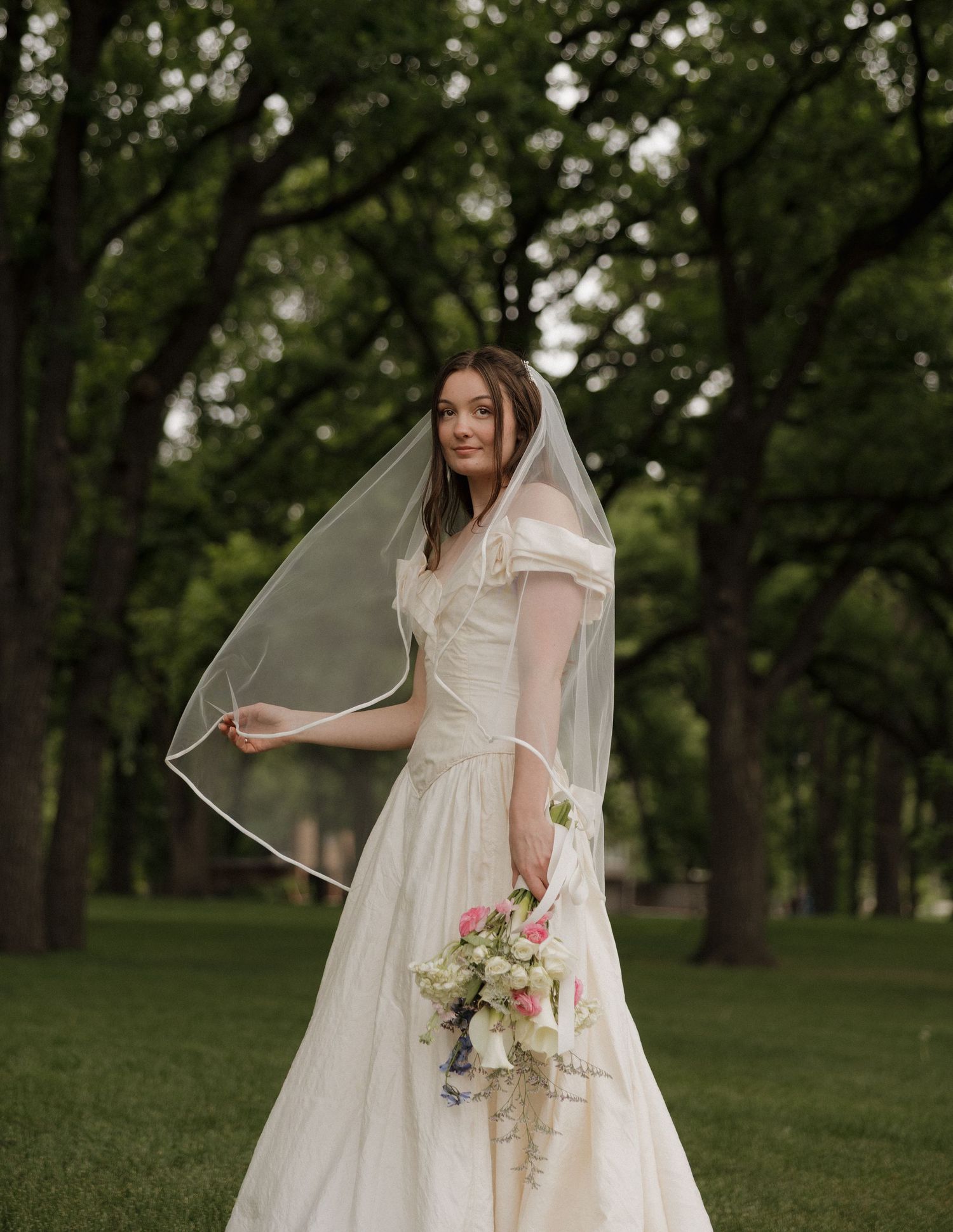 Bride in white gown and veil holds floral bouquet outdoors among lush green trees on a cloudy day.