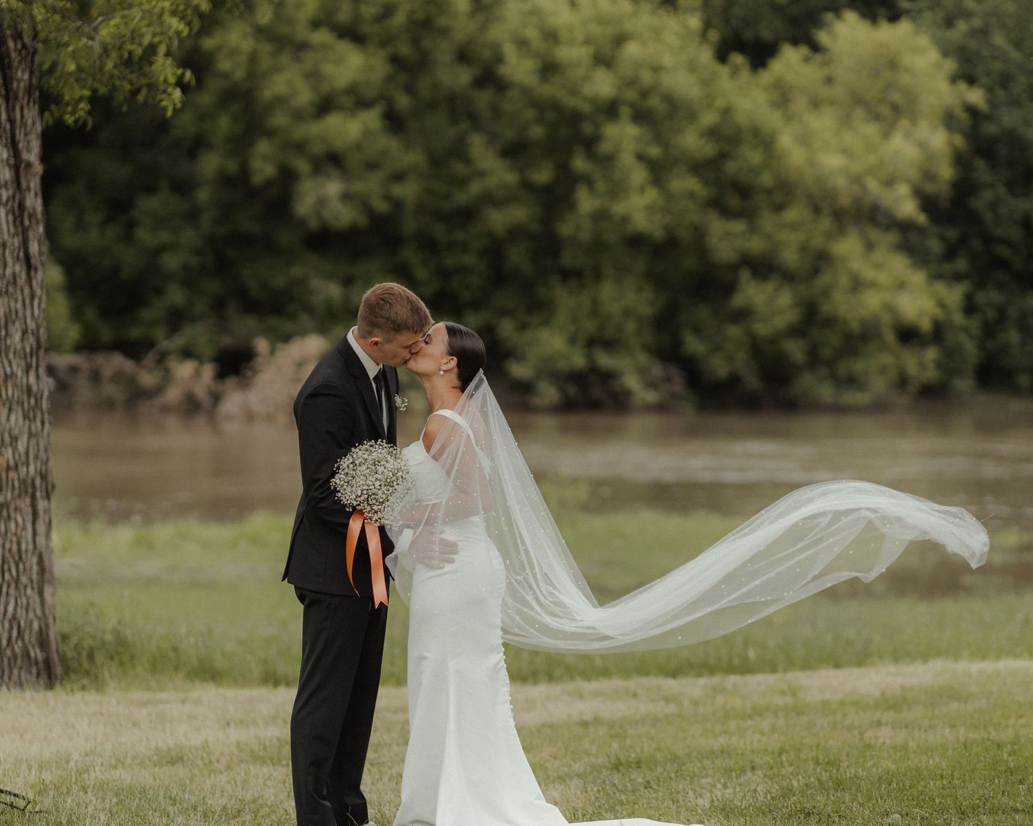A wedding couple stands in a grassy field while a long white veil flows gracefully in the breeze.