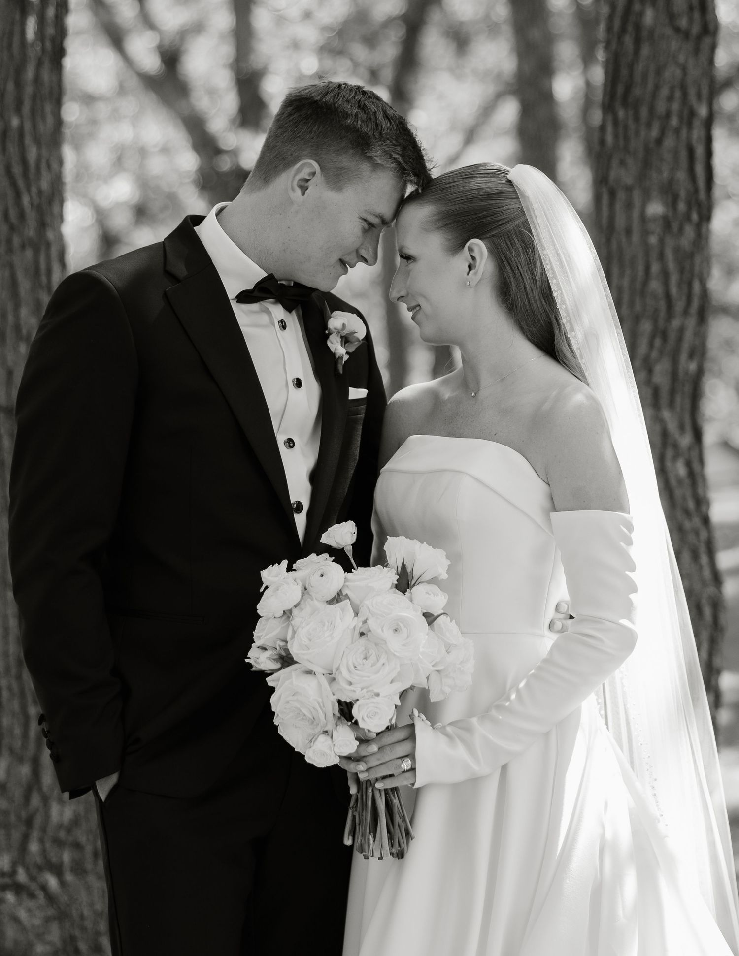 A romantic black and white wedding portrait series showing a couple in formal attire embracing among tall trees.