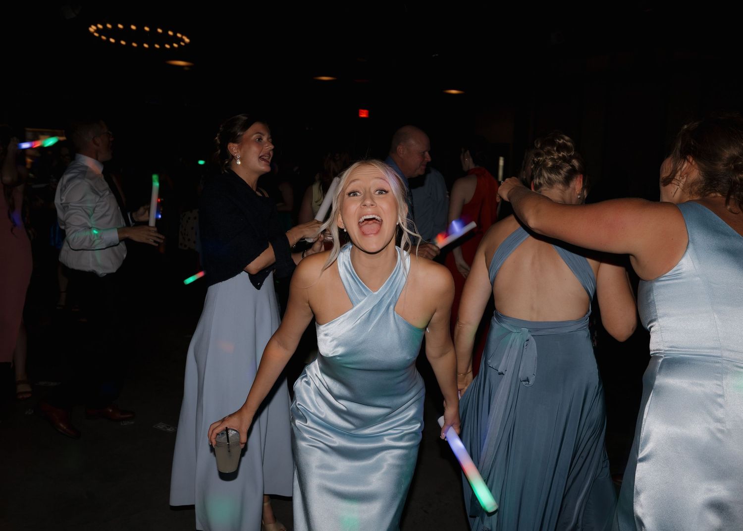 Dancers in light blue dresses celebrate on a dark dance floor with glow sticks at an evening party.