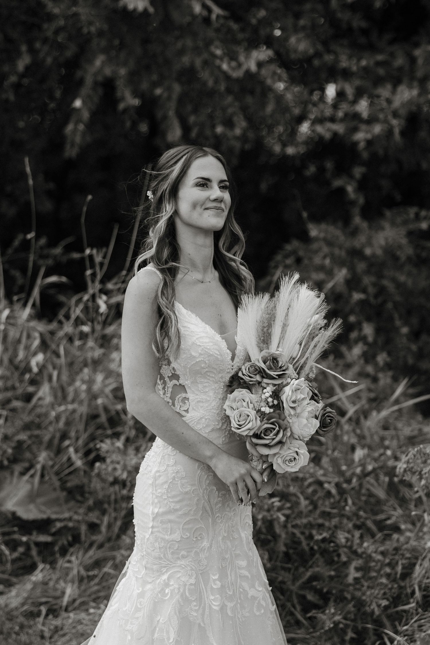 A bride in a white lace wedding dress holds a bouquet with pampas grass and roses in an outdoor garden setting.