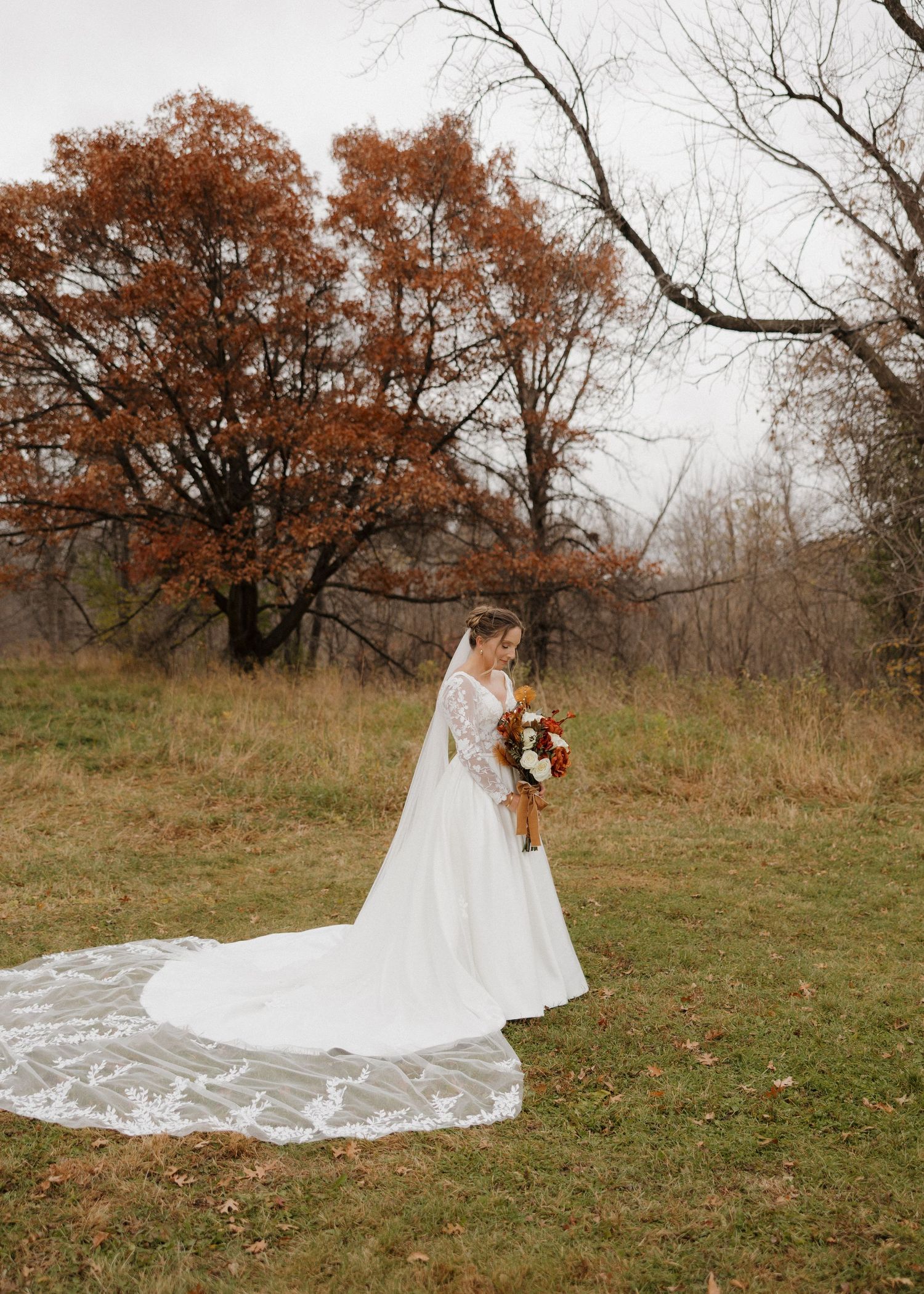 A long white wedding dress train flows gracefully across an autumn field with bare trees in the background.