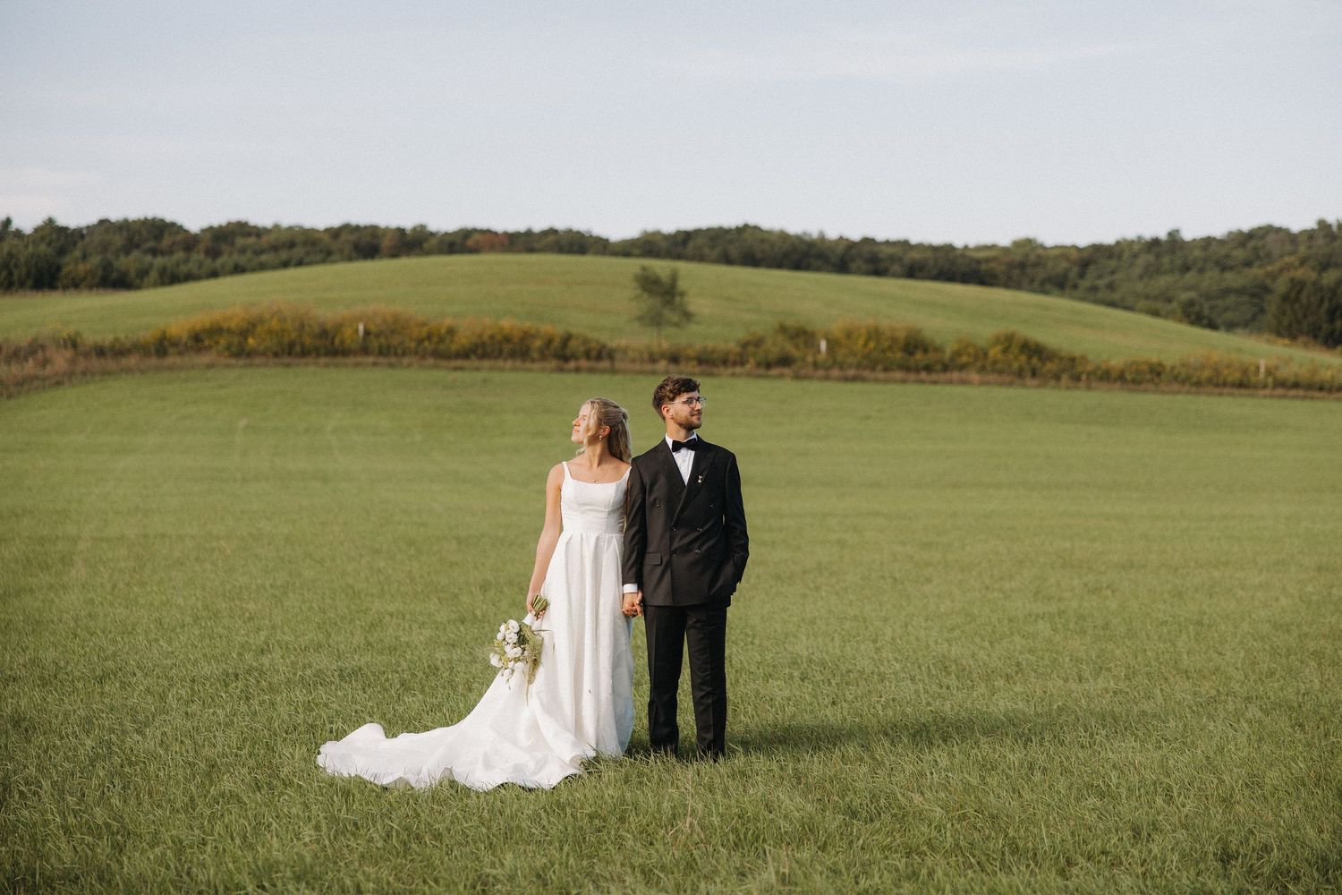 A couple in wedding attire stands together in a scenic green hillside meadow during their special day.