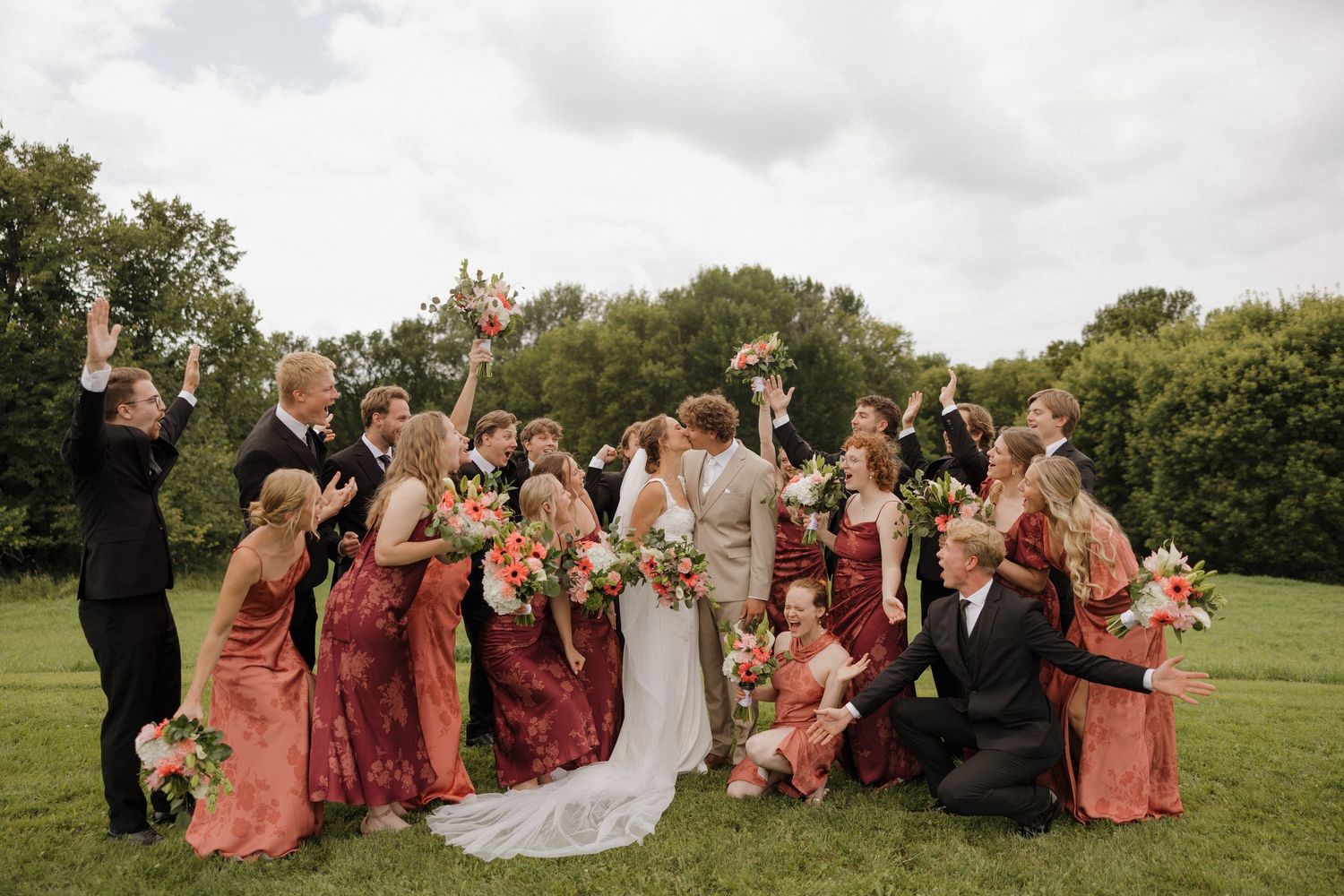 Wedding party poses together on a grassy lawn in burgundy and coral attire during an outdoor celebration.