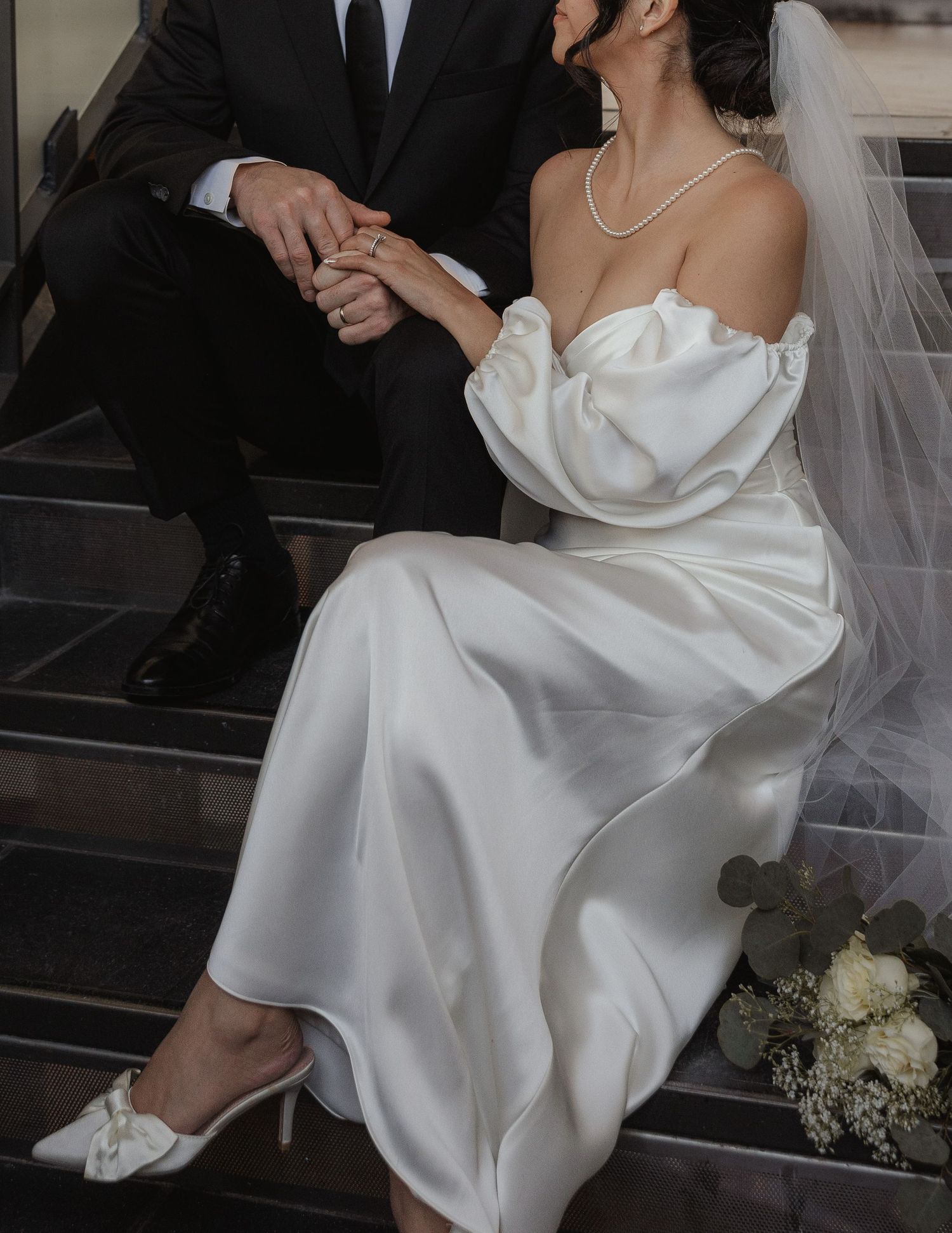 Bride in elegant white wedding dress sits gracefully on dark stairs while holding her flowing gown.
