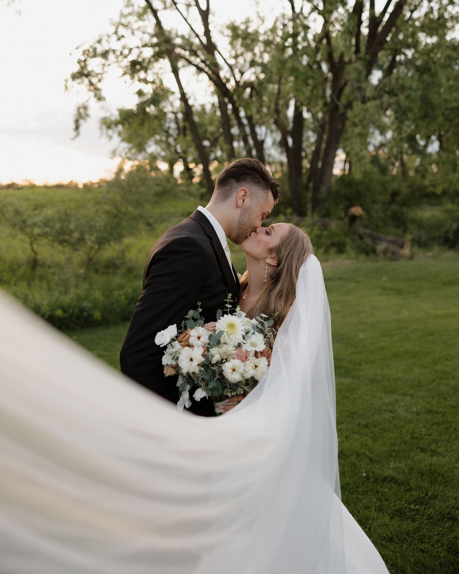 A bride and groom share a romantic kiss outdoors with a flowing white veil trailing behind them.