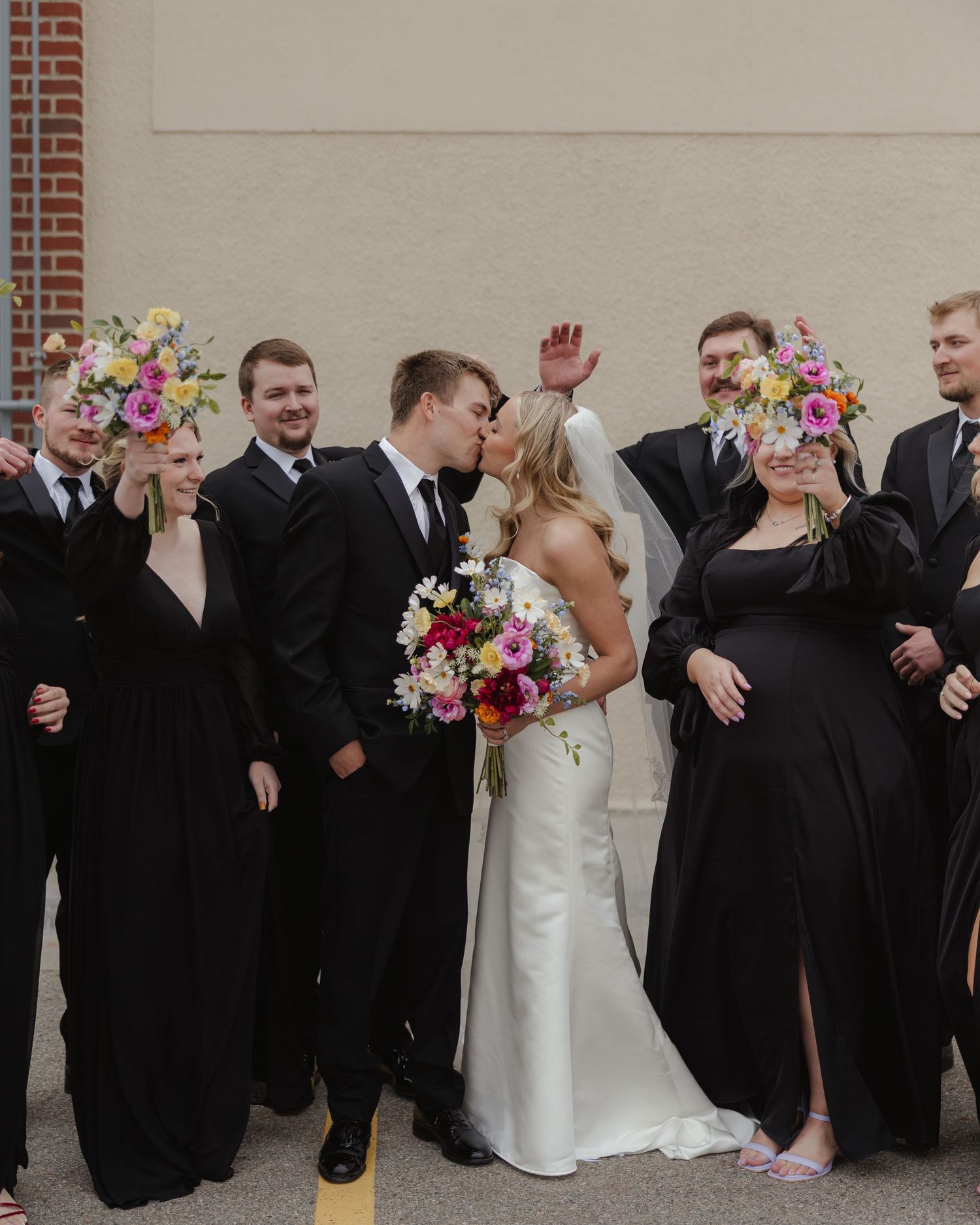 Wedding party members in formal black attire and white gowns celebrate joyfully in a line against a brick wall.