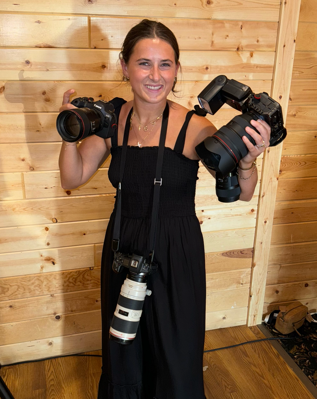 A photographer poses with multiple professional cameras in a wooden-paneled room.