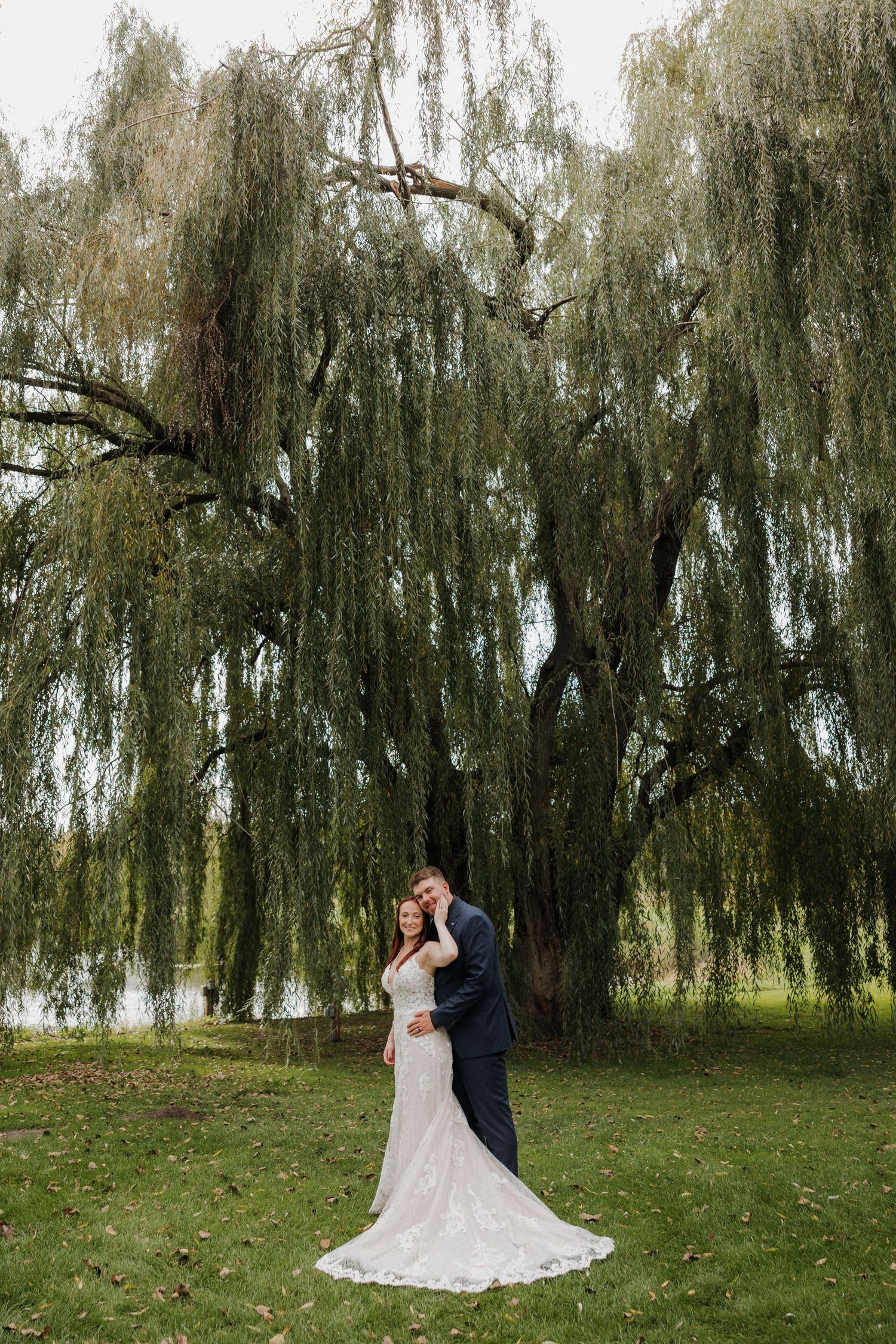 Romantic wedding couple poses beneath drooping willow trees on a lush green lawn.