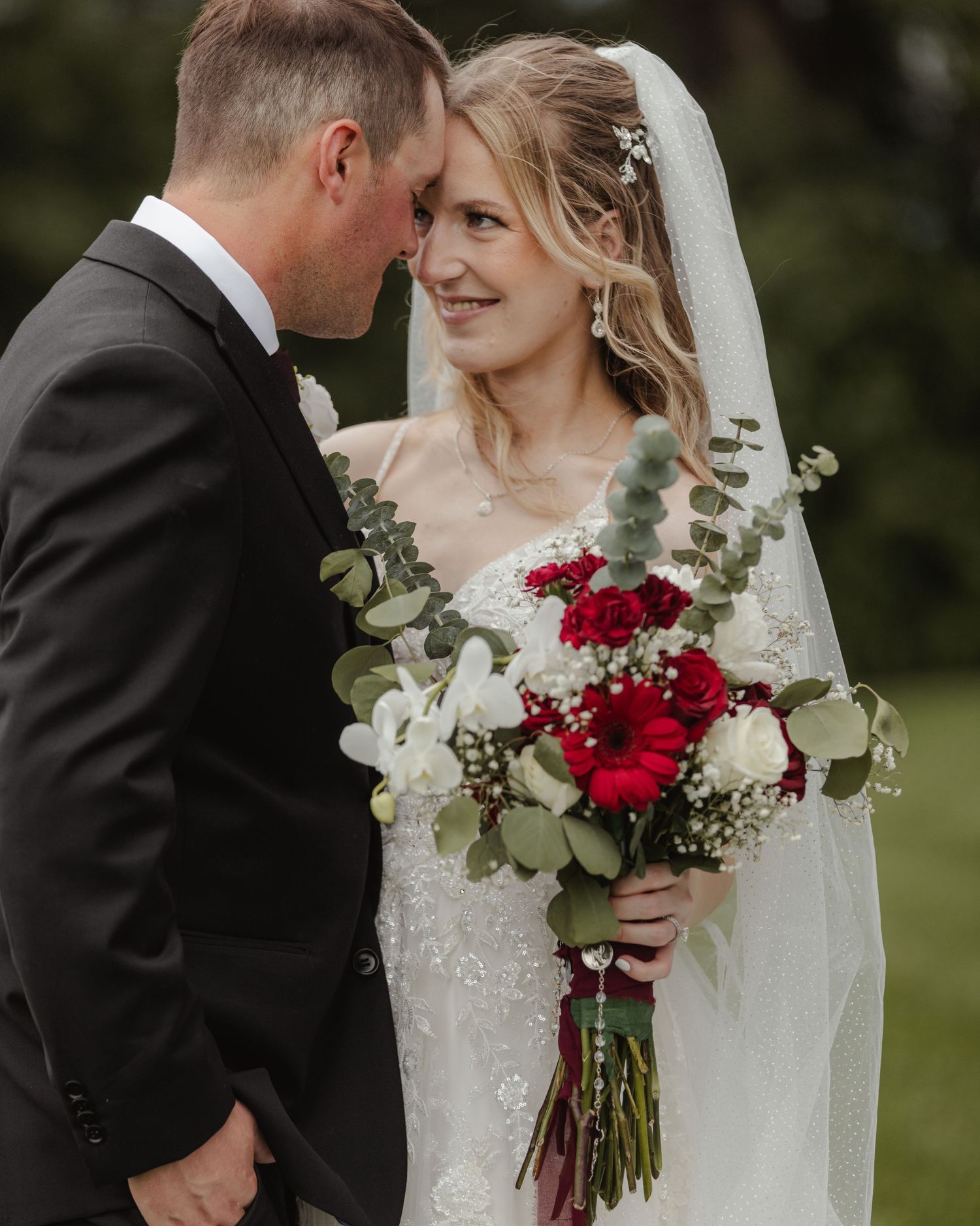 A couple shares a romantic moment at their wedding with red and white bouquet against a natural outdoor backdrop.