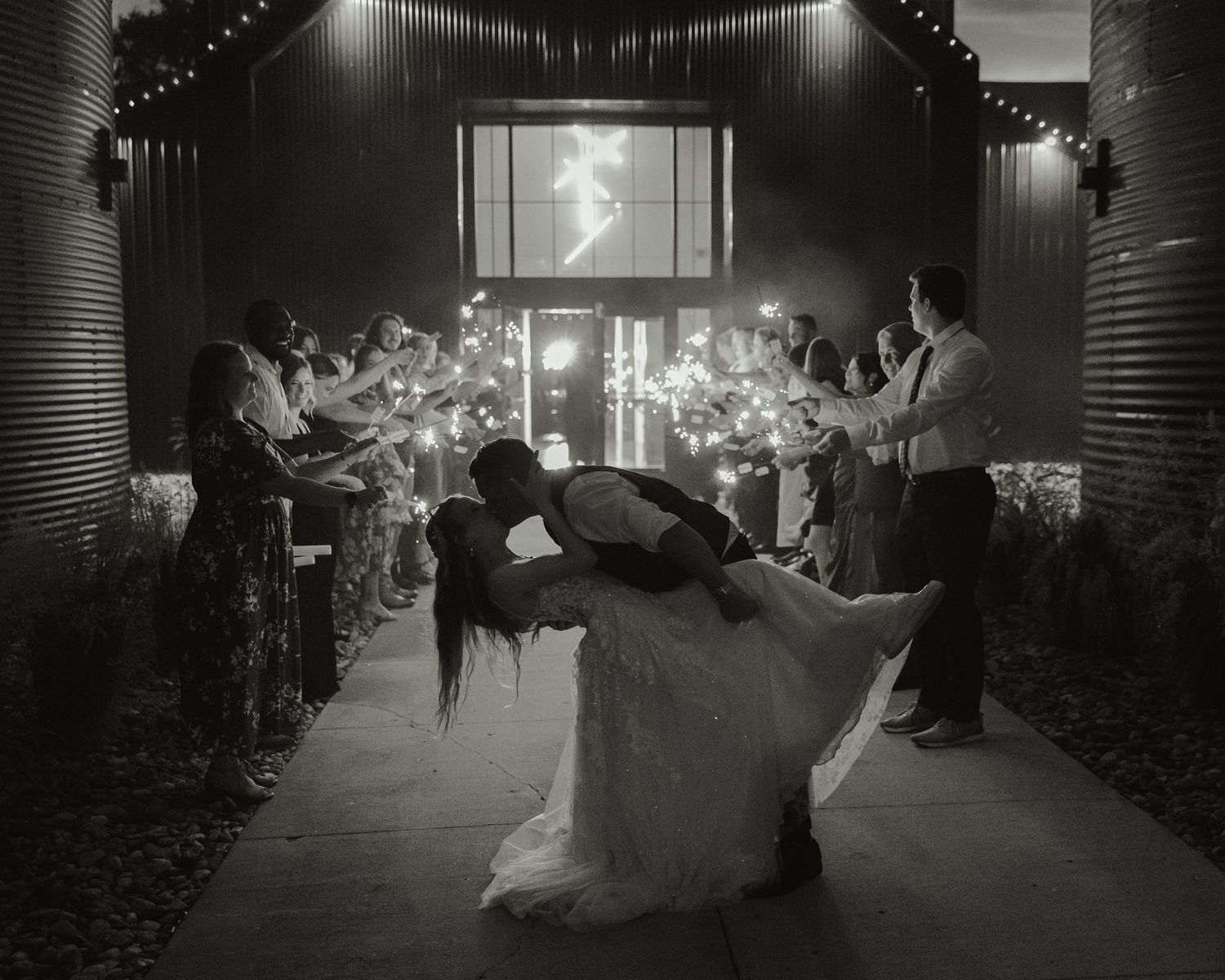 A dramatic black and white wedding scene shows a couple dancing in an illuminated corridor with columns.