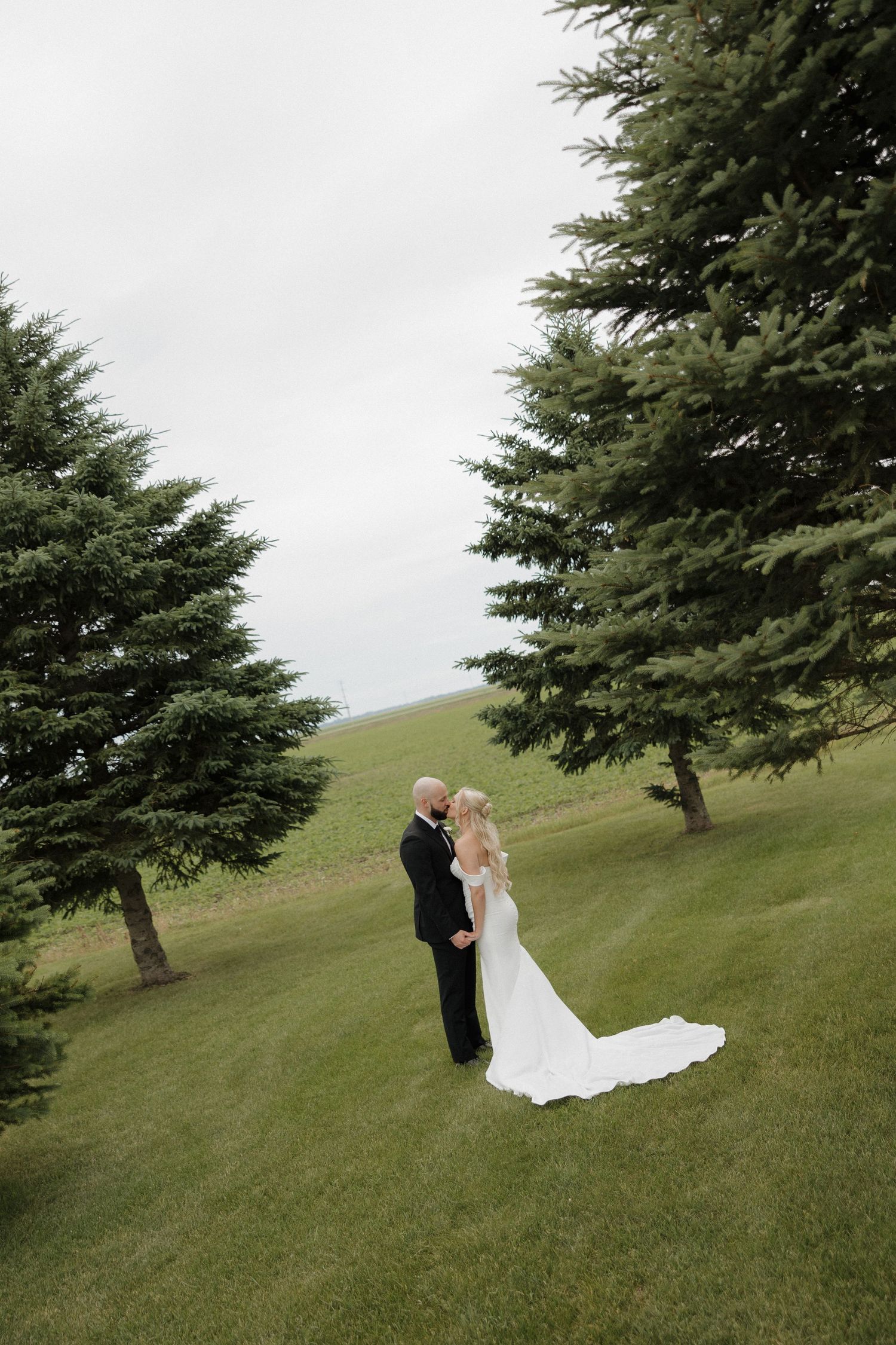 A bride and groom share a romantic moment on a lush green hillside surrounded by tall evergreen trees on their wedding day.