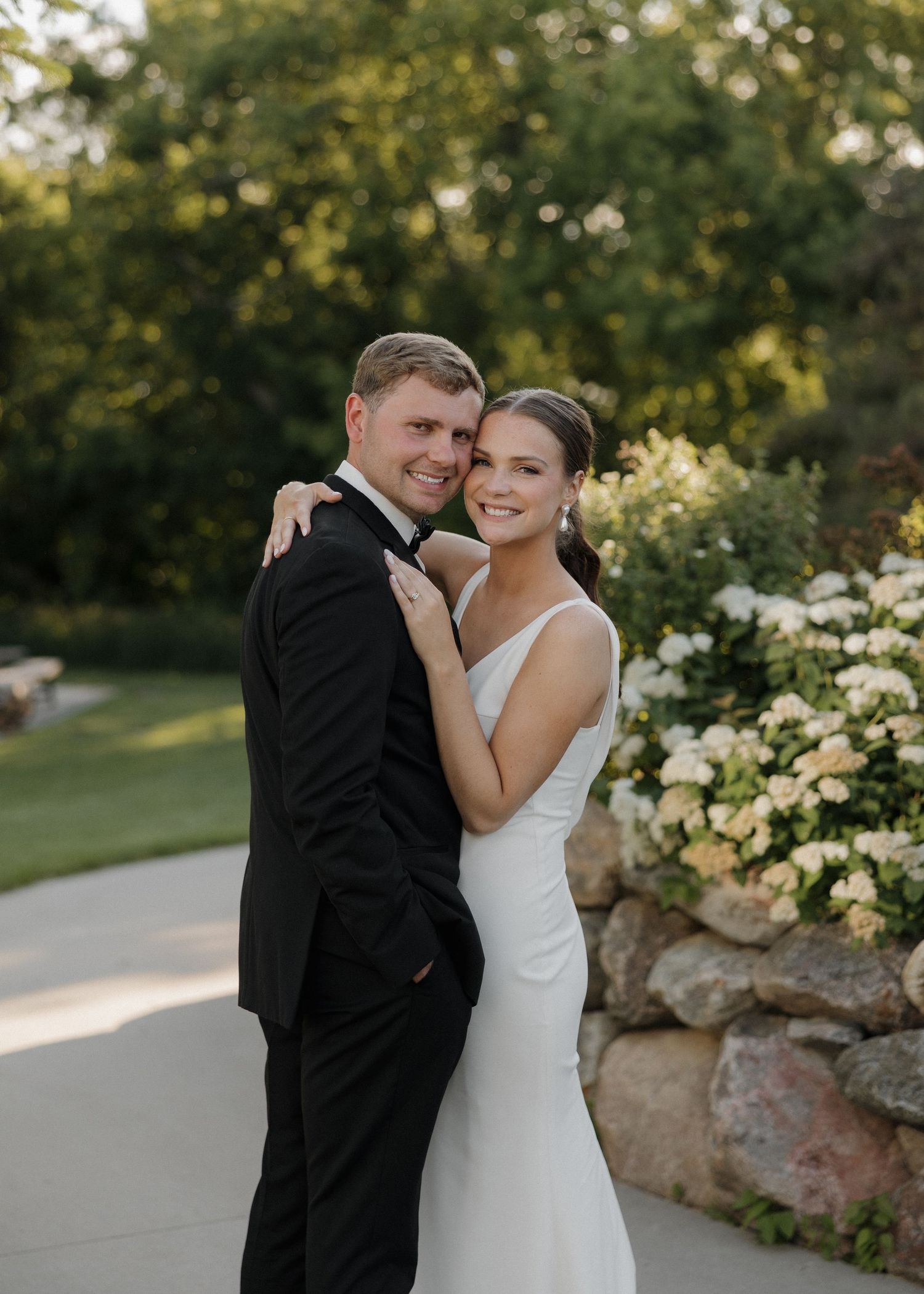 Bride and groom pose together outdoors near floral arrangements, groom in black tuxedo, bride in white gown.