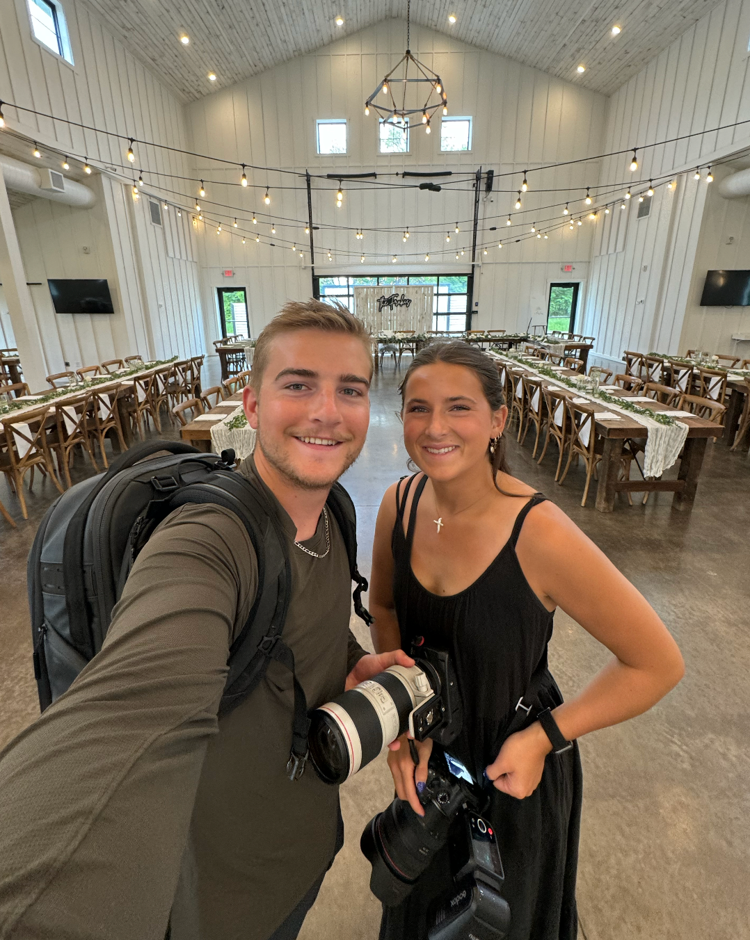 Two photographers with professional cameras take a selfie in a rustic wedding venue with string lights.