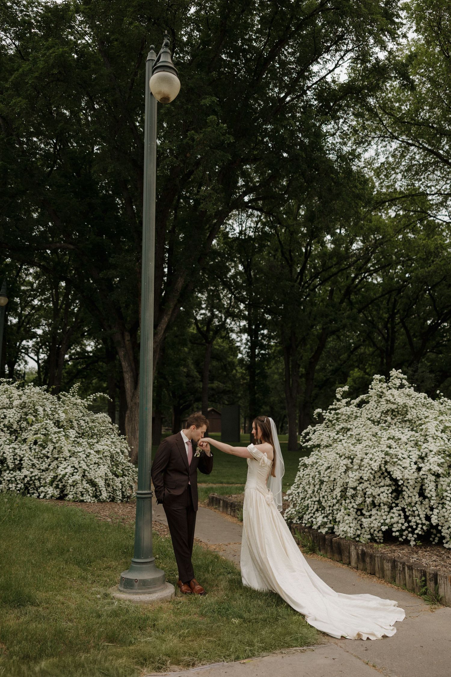 A bride and groom share a romantic moment near a lamppost surrounded by blooming white bushes in a lush green park.