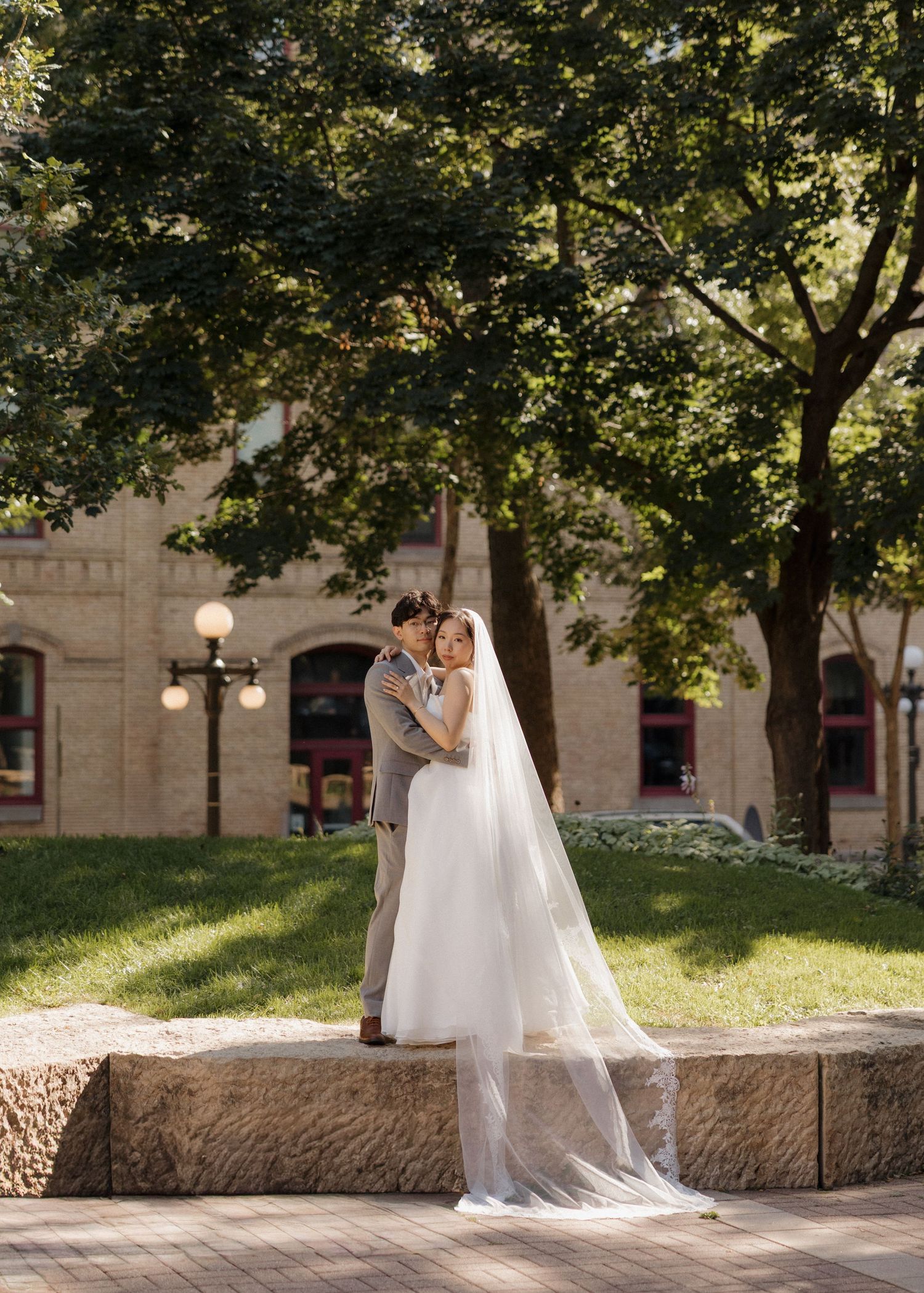 A bride in a flowing white gown stands gracefully on stone steps in a sunlit garden setting with trees in the background.