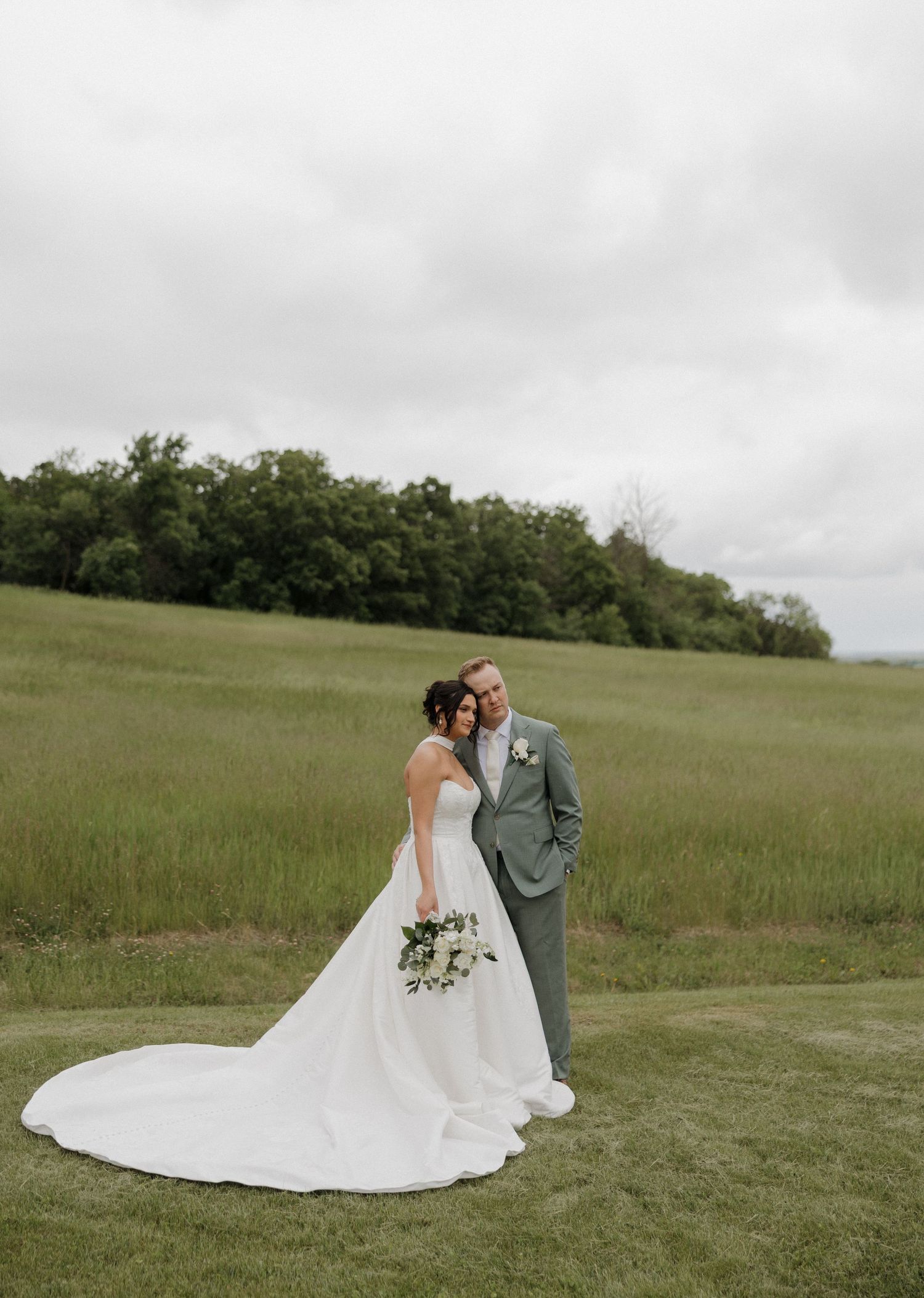 A bride and groom pose in a lush green meadow, the bride in a flowing white gown holding a bouquet on their wedding day.