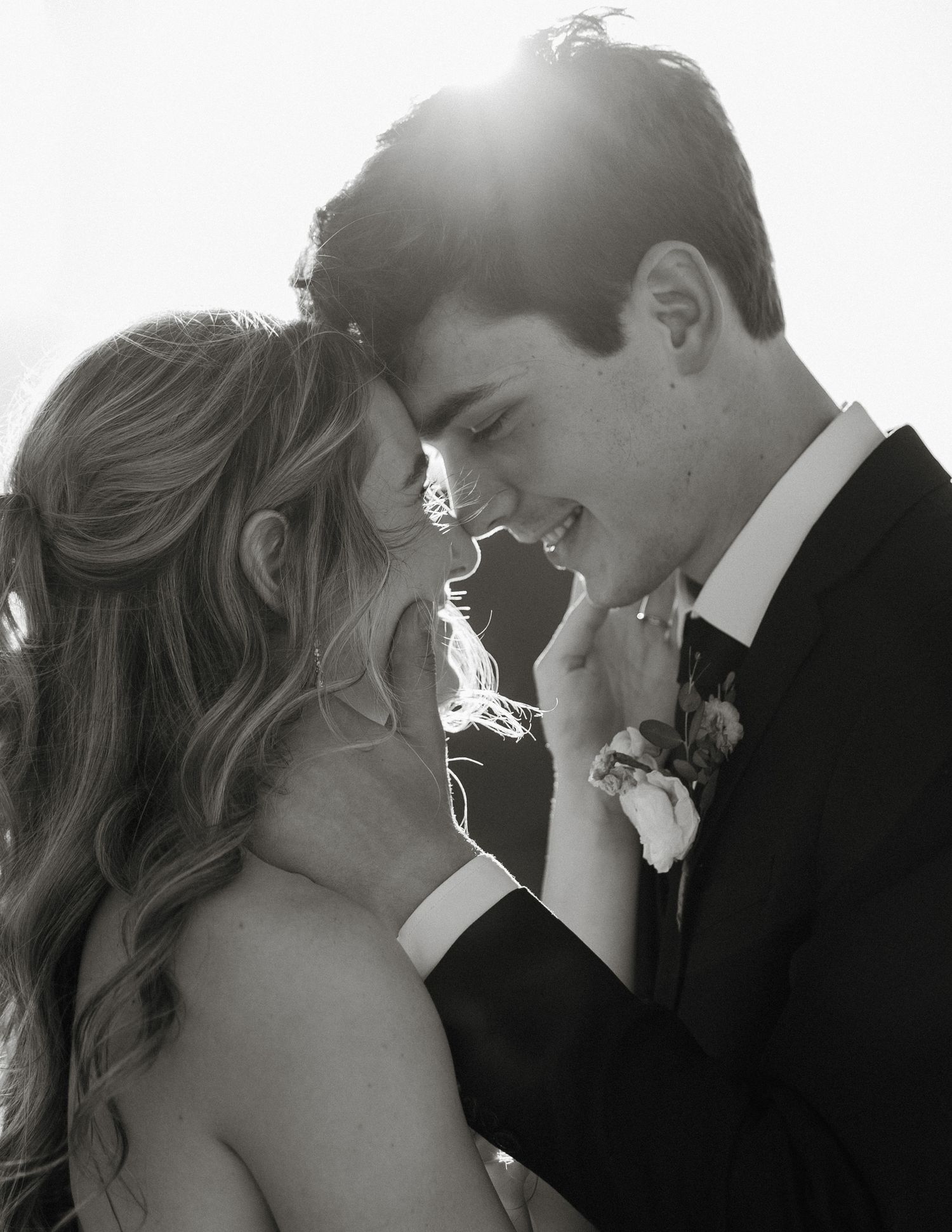 Black and white wedding photo of a smiling couple in formal attire sharing an intimate moment with sunlight glowing behind them.