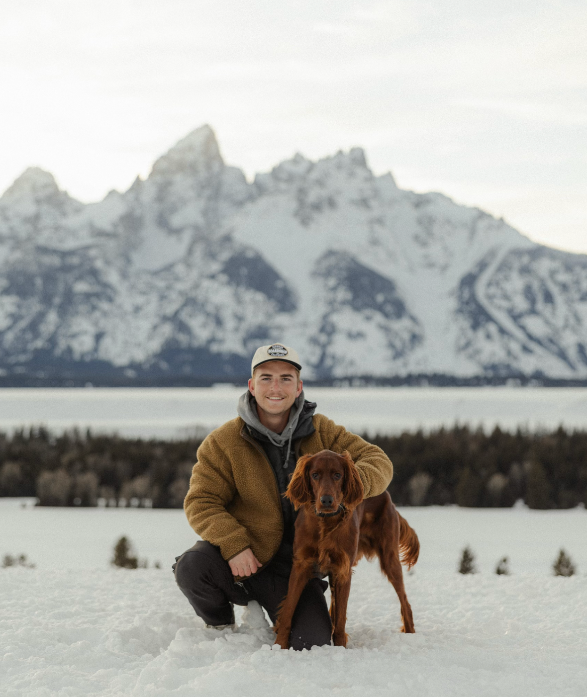 A person poses with their red retriever dog in front of snow-capped mountains.