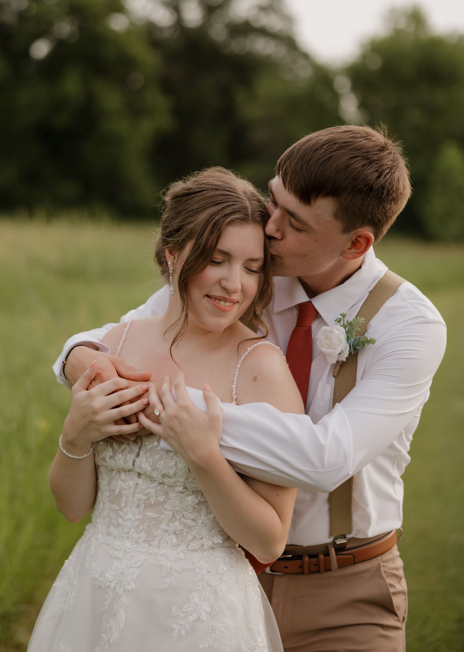 A romantic couple shares tender moments in an outdoor field setting with the bride in a white dress and groom in suspenders.