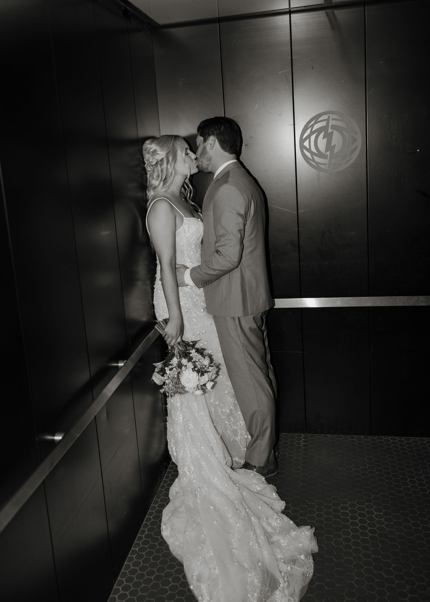 Black and white photo of a bride and groom sharing an intimate moment in an elevator, with her lace dress trailing behind.