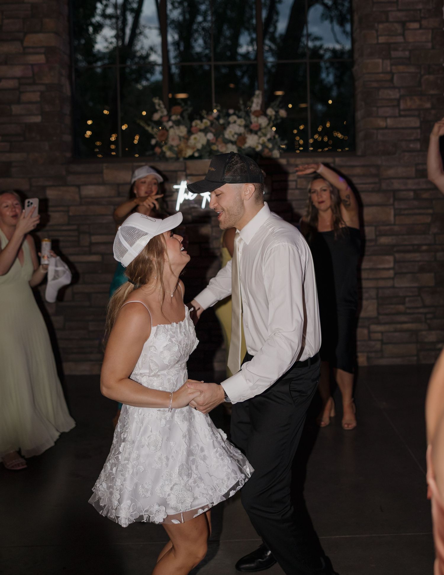 Wedding guests dance and celebrate together during an evening reception at a brick-walled venue.