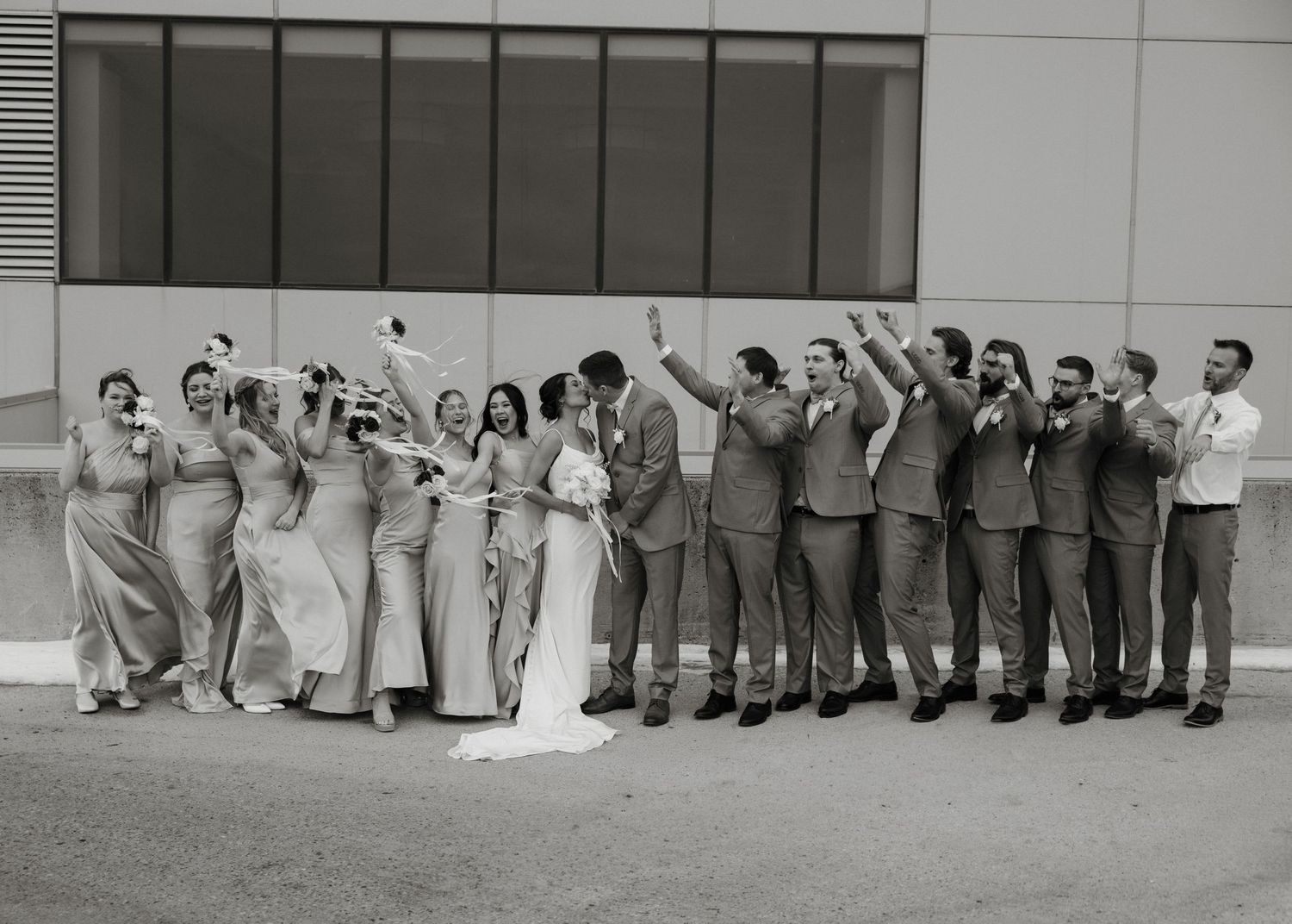 A wedding party in formal attire stands in a line outside, with bridesmaids in gray dresses and groomsmen in suits.