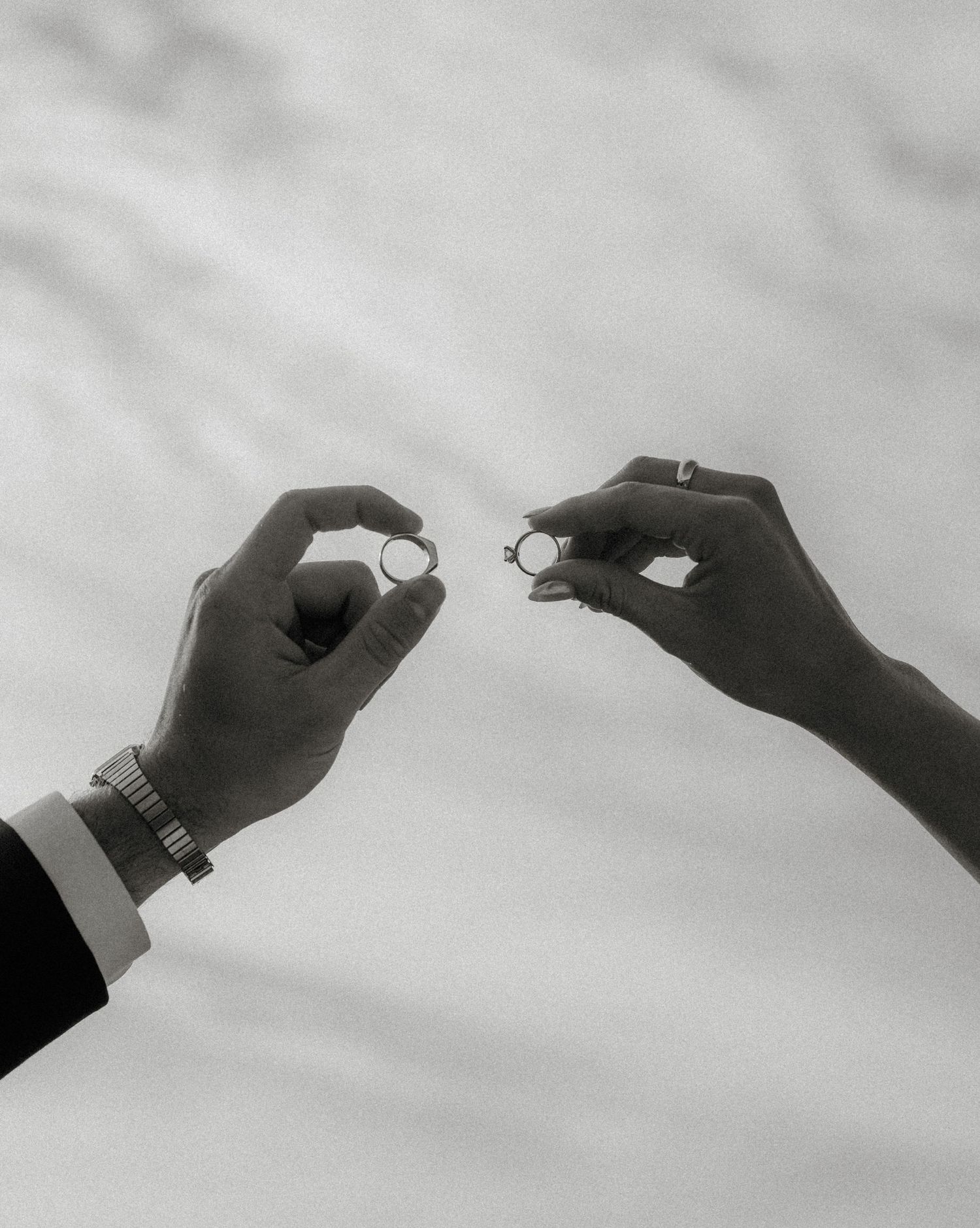 Black and white photo of two hands reaching towards each other holding wedding rings against a cloudy sky.
