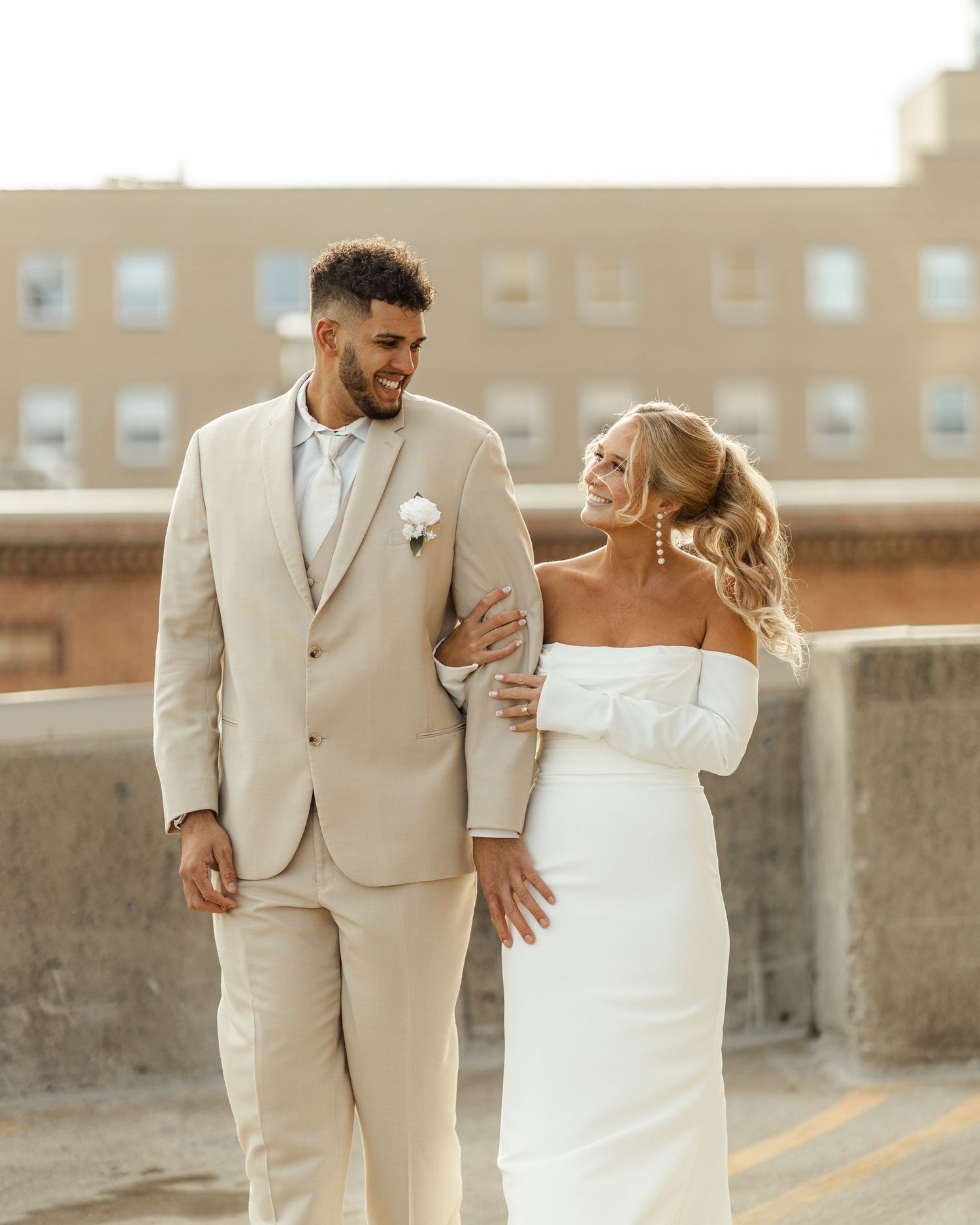 A couple in wedding attire share a tender moment while walking along a sunlit brick wall outdoors.