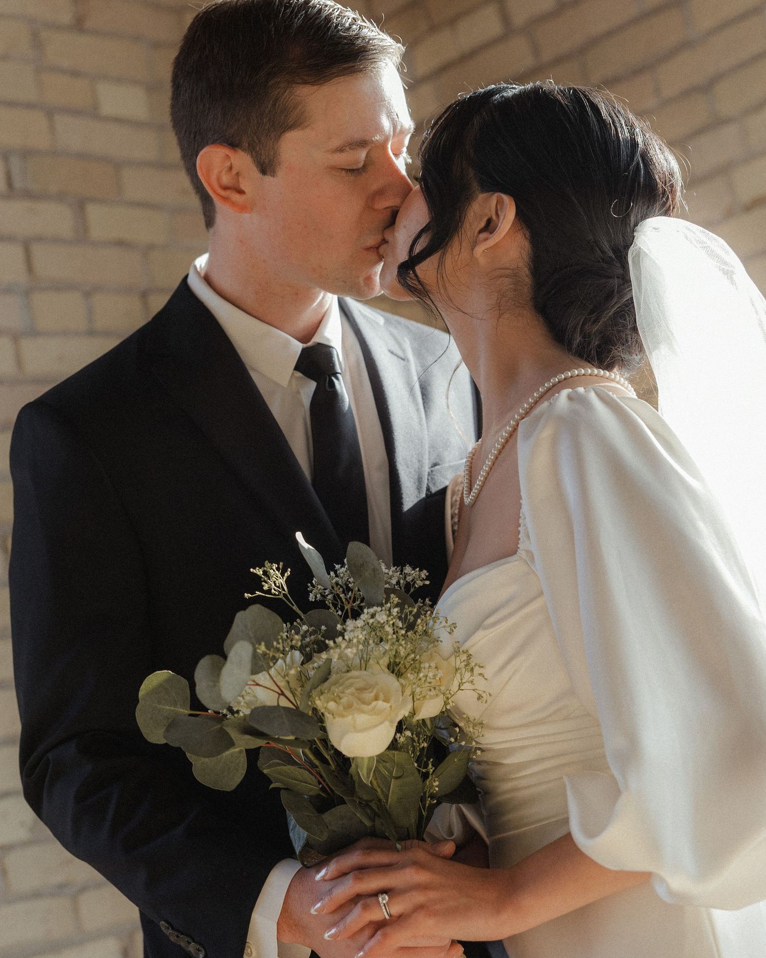 A romantic wedding couple shares tender moments in four sequential photos against a brick wall with white flowers.
