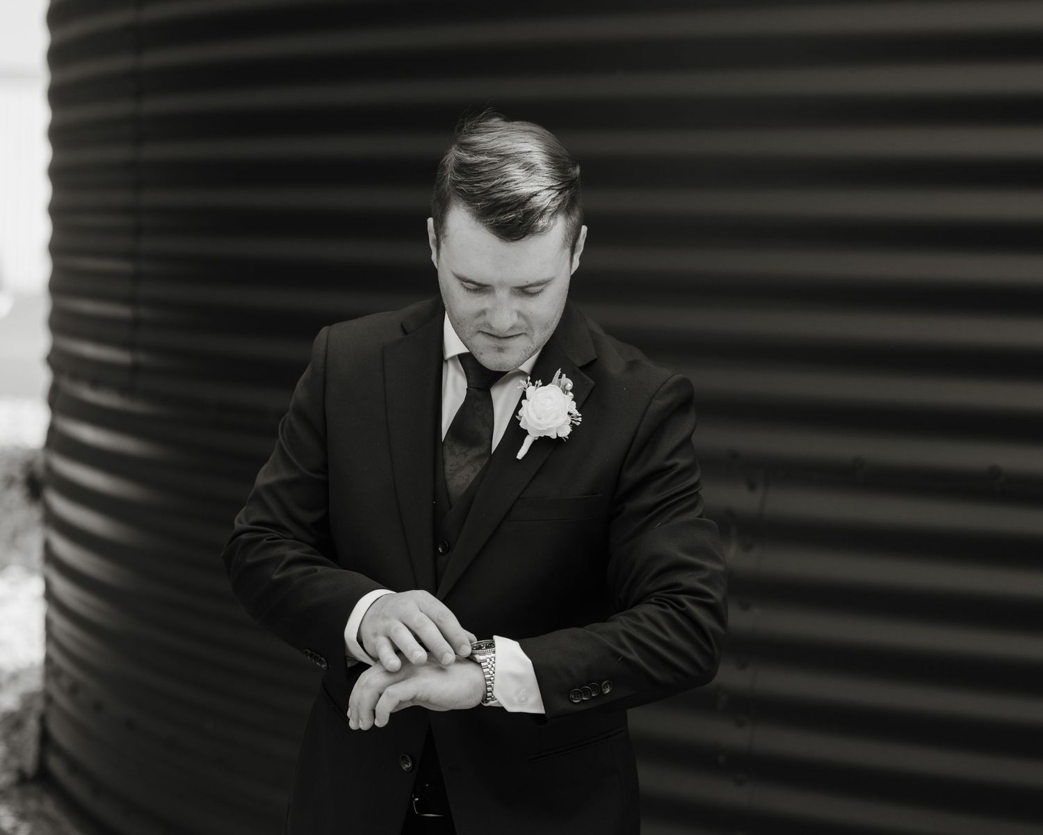 A person in a black suit and white shirt stands against a dark striped background in a black and white photograph.
