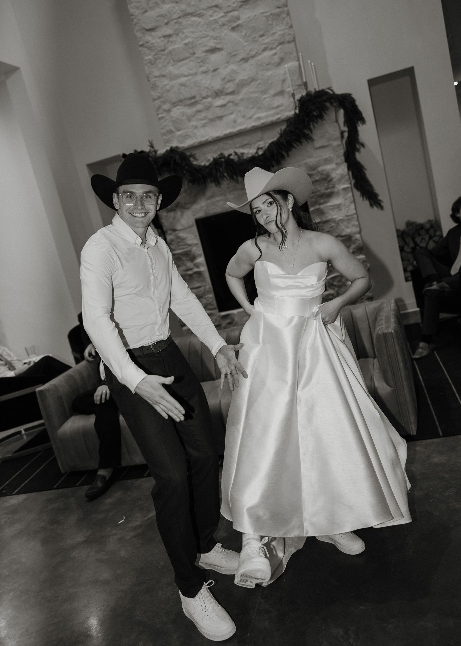 A black and white photo of a couple in western attire with cowboy hats posing together in a rustic setting.