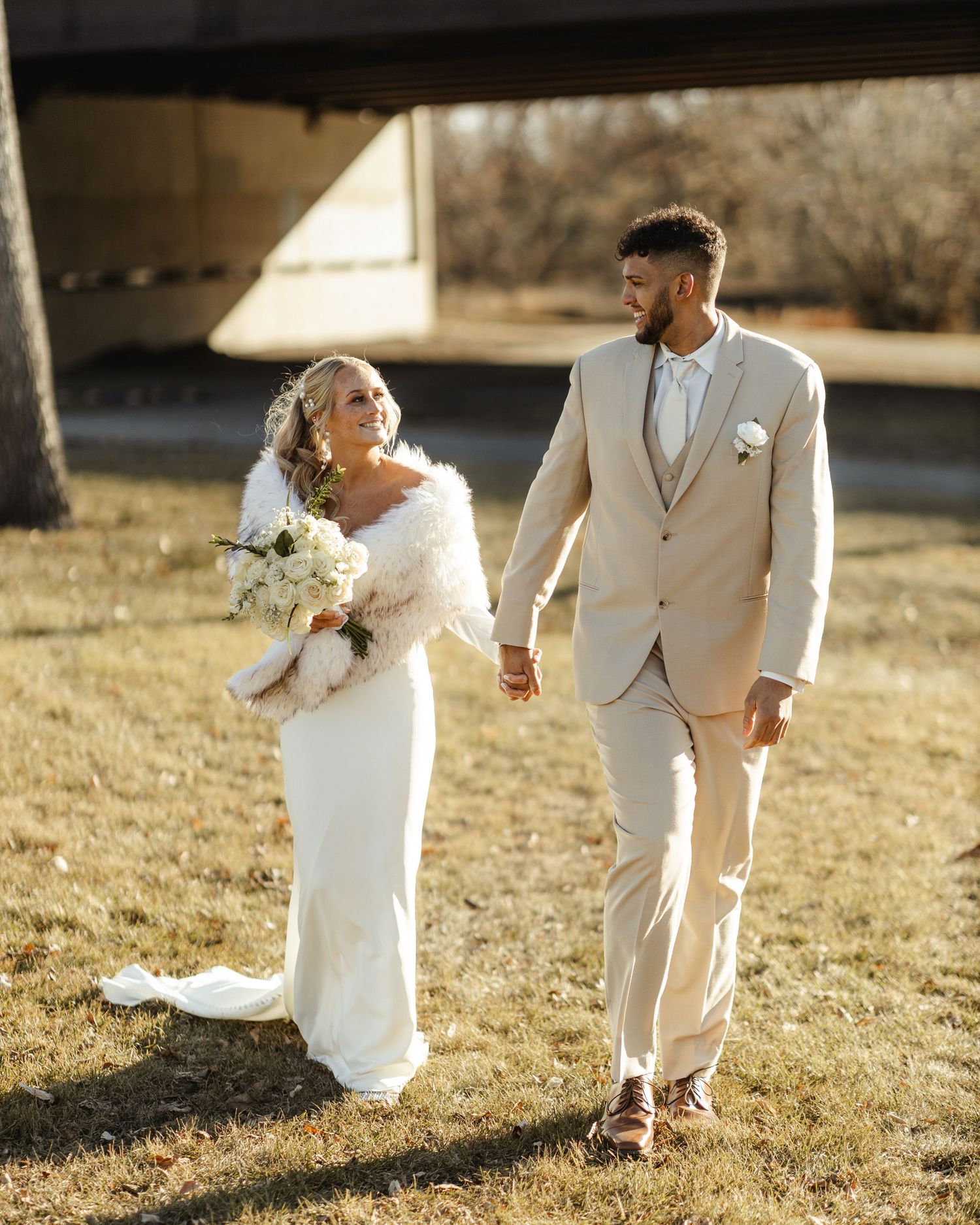 A couple in white wedding attire walks through a sunlit field during golden hour.