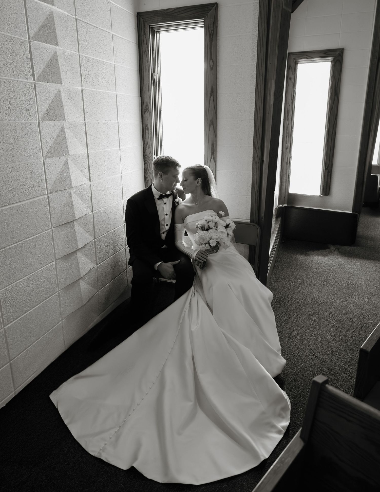 A romantic black and white wedding photo showing a couple in formal attire sharing an intimate moment by a window.